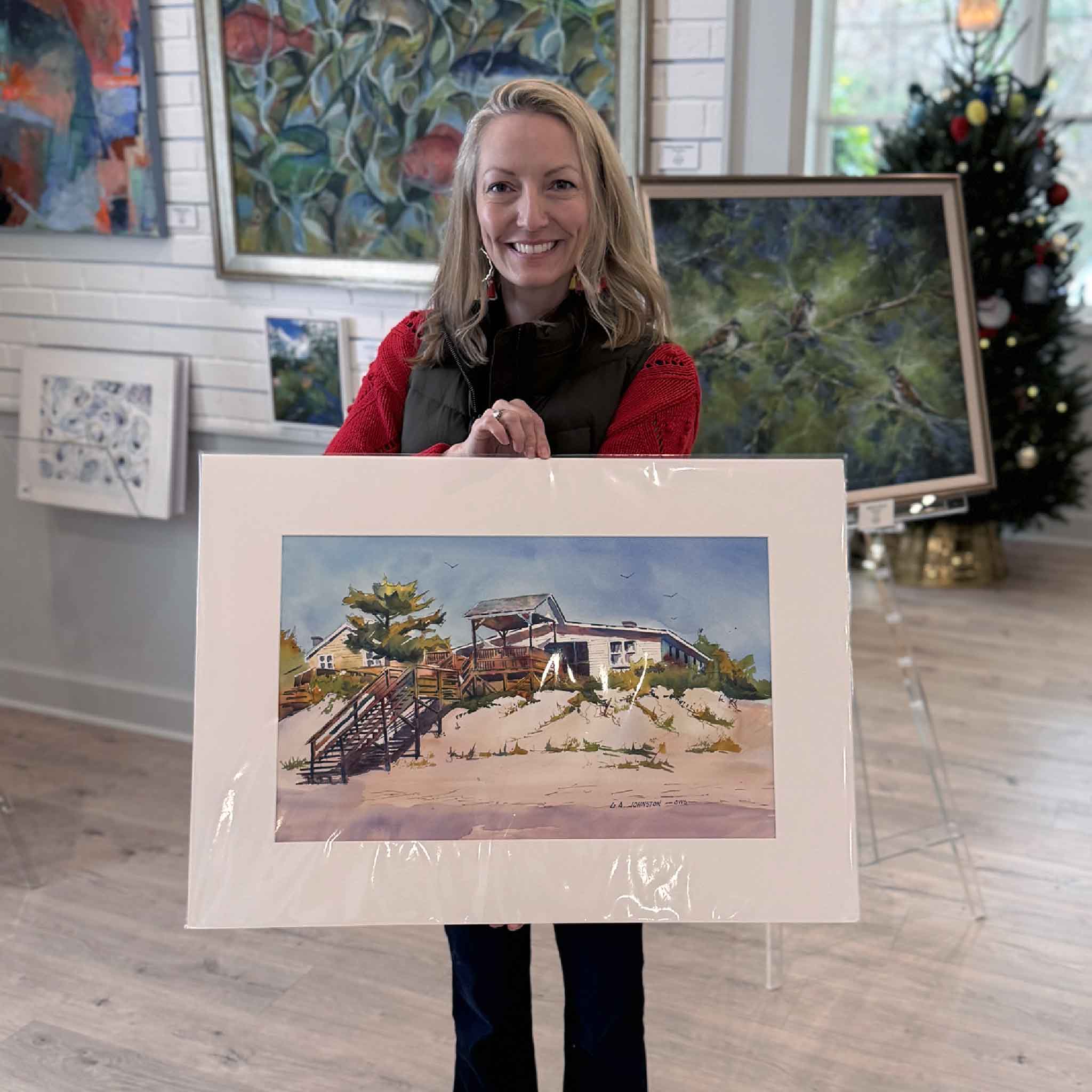 Local artist Jeanne O'Neal standing inside Gray Man Gallery holding the matted Rowell’s Retreat watercolor by Gary Johnston.