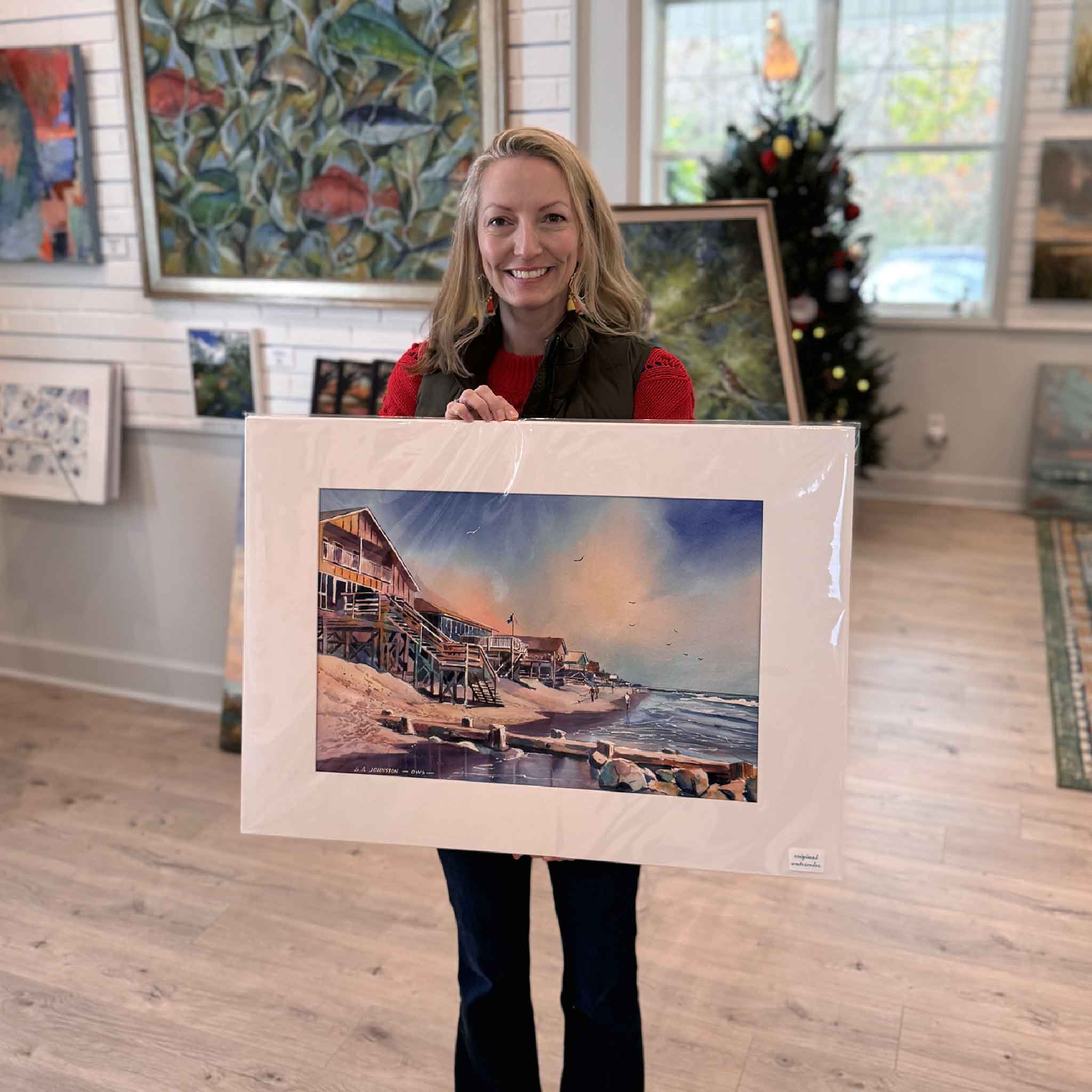 Local artist Jeanne O'Neal holds the matted South End watercolor by Gary Johnston inside Gray Man Gallery, showing the artwork’s scale and vibrant coastal detail.