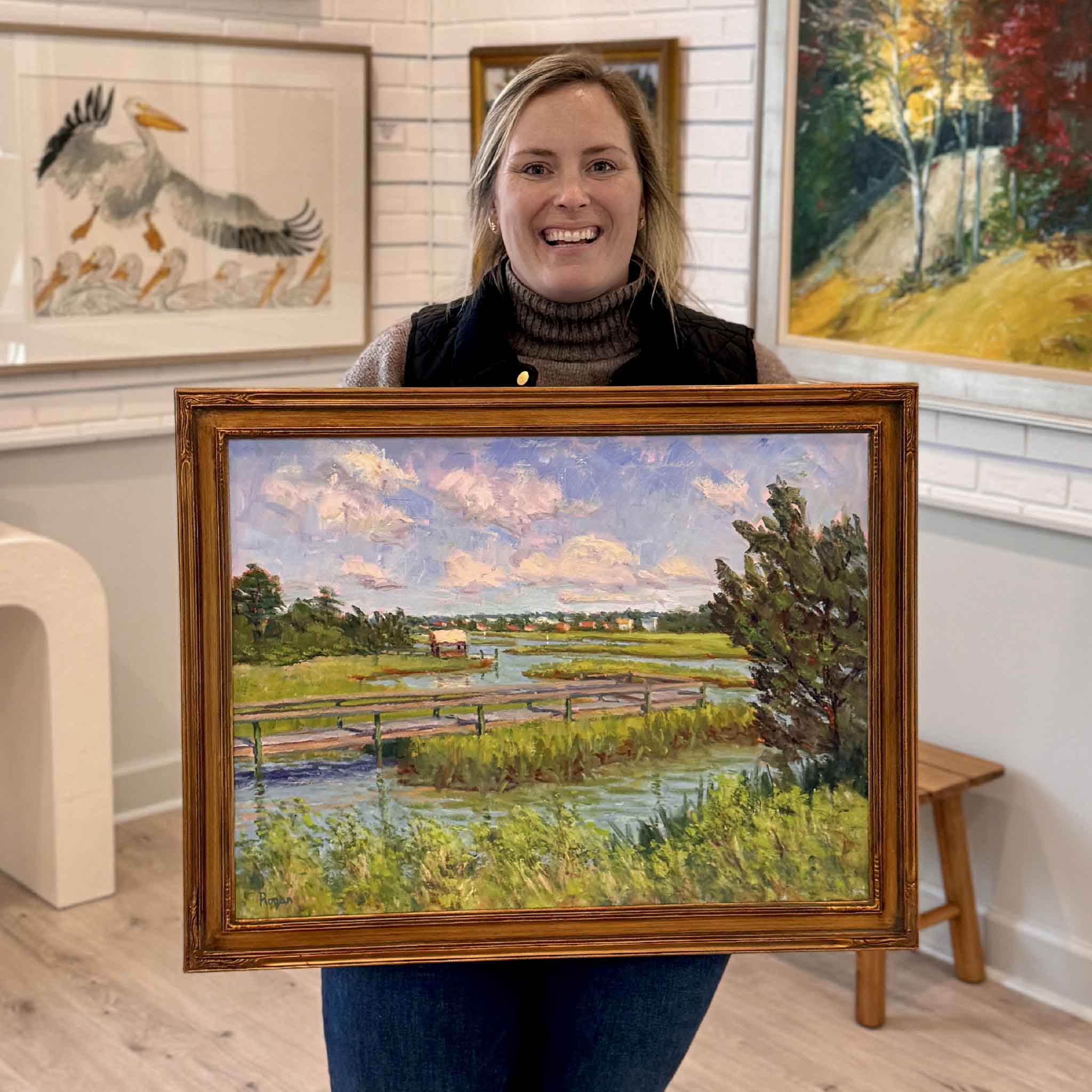 Smiling Gray Man Gallery staff member holding “South Litchfield View,” an original oil painting by local artist Michael Rogan, featuring Lowcountry marshes, tidal creek, and bridge near Pawleys Island, South Carolina, displayed inside Gray Man Gallery