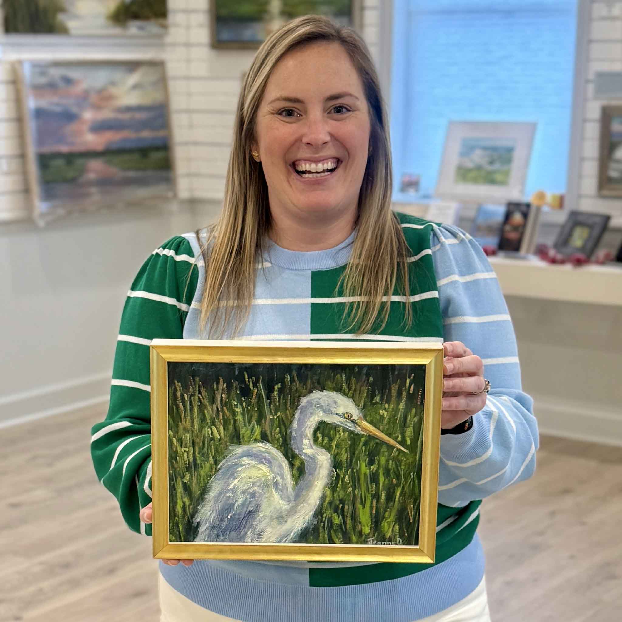 Gray Man Gallery team member holding Sun Dappled: A Study by local artist Jeanne O’Neal, original oil painting of egret in marsh, photographed inside Pawleys Island gallery