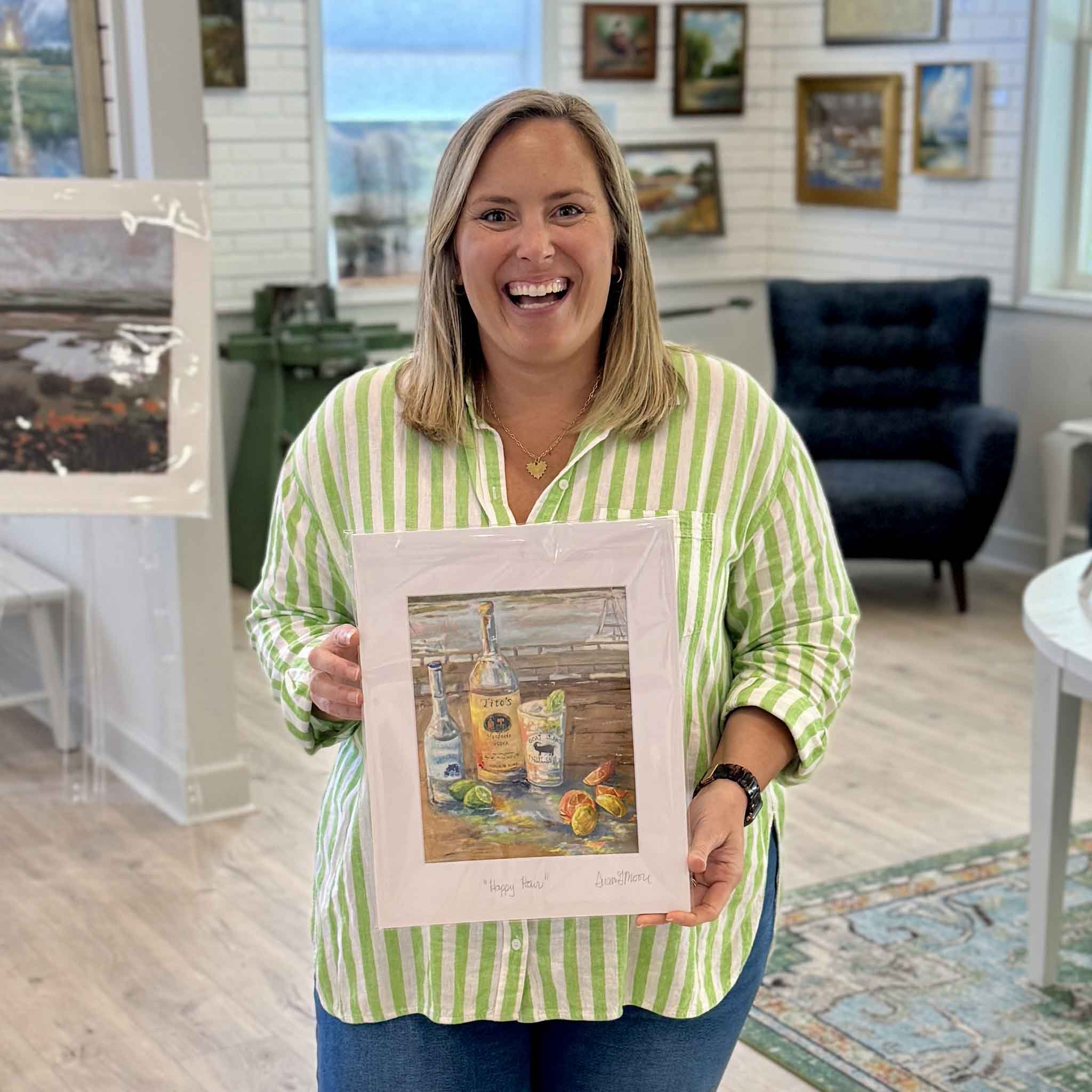 Team member Bordy holding Susan Moore Happy Hour fine art giclee print matted to 11x14 showing coastal still life with Tito’s bottle, citrus, and cocktail on a dock, available at Gray Man Gallery in Pawleys Island