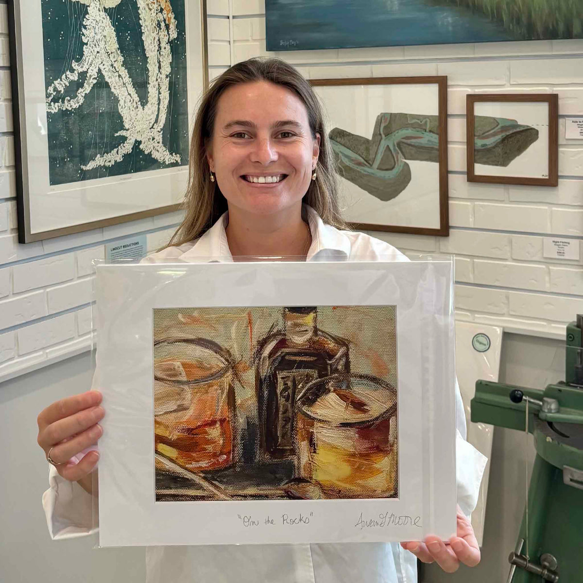 Kyra holds On the Rocks by Susan Moore at Gray Man Gallery in Pawleys Island, South Carolina, a playful whiskey and cocktail fine art print featuring two amber drinks and a bottle, matted to a standard 11 x 14 size for easy framing.