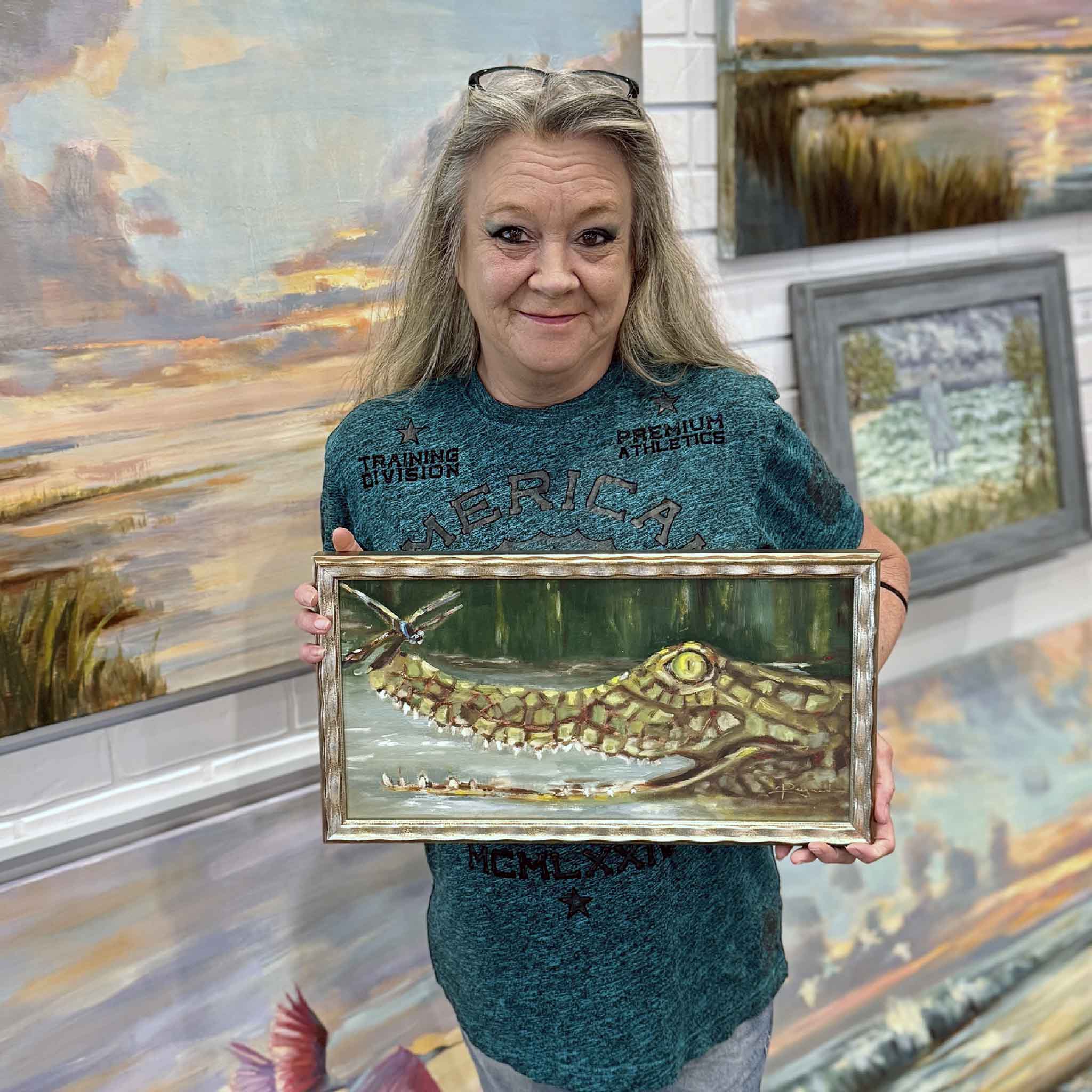A Gray Man Gallery team member holds the framed Swamp Thing original oil painting by Jenny Reynolds, showing the scale of the whimsical marsh gator artwork.