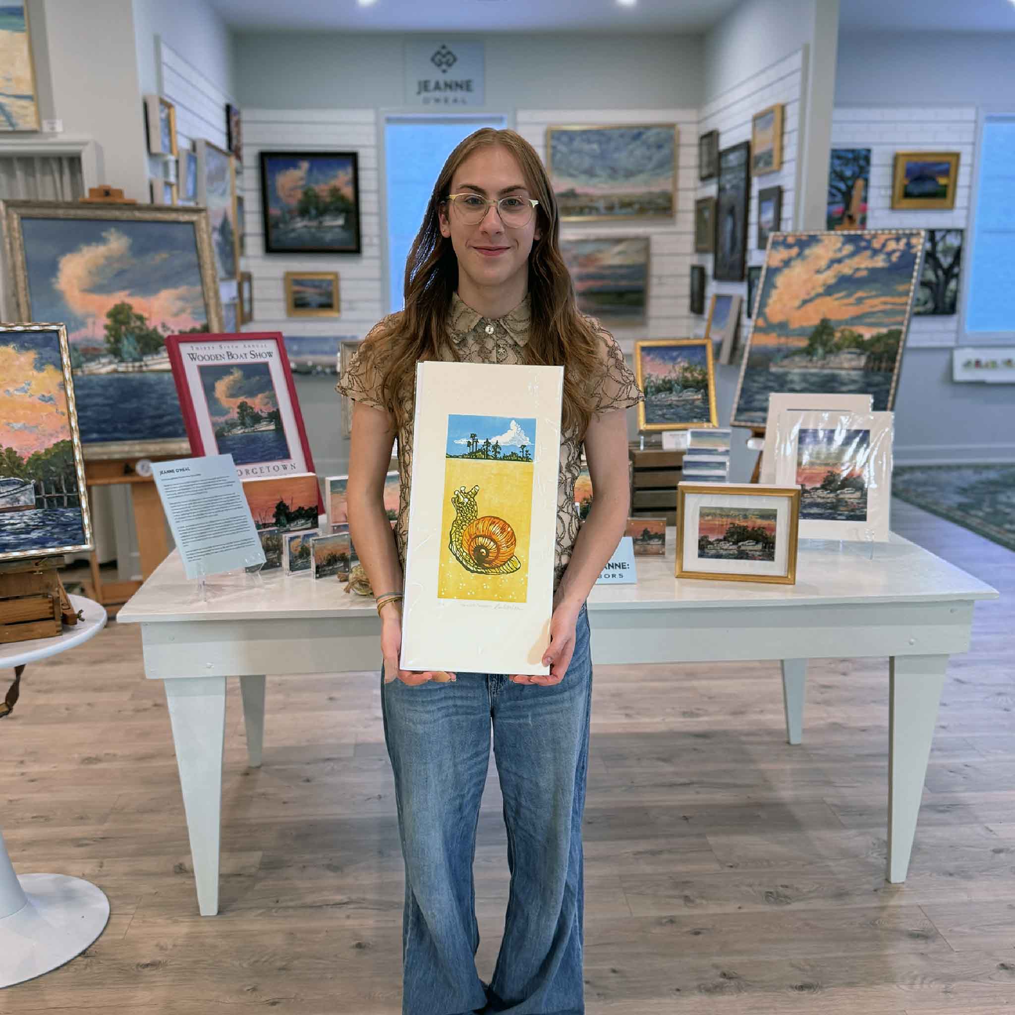 Alex holding “The Long Journey Home,” an original linocut by Laurie McIntosh featuring a coastal scene with a shell and palm trees, displayed inside Gray Man Gallery.