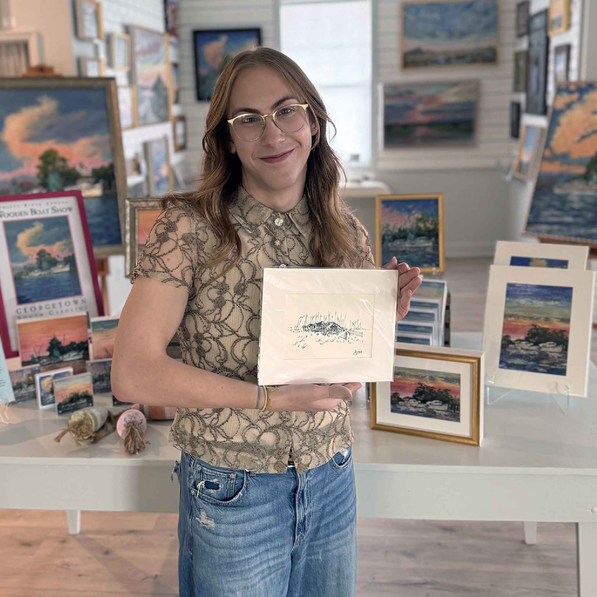 Alex holding the matted ‘Beach House with Sea Oats #11’ coastal sketch by Todd Halamka inside Gray Man Gallery.