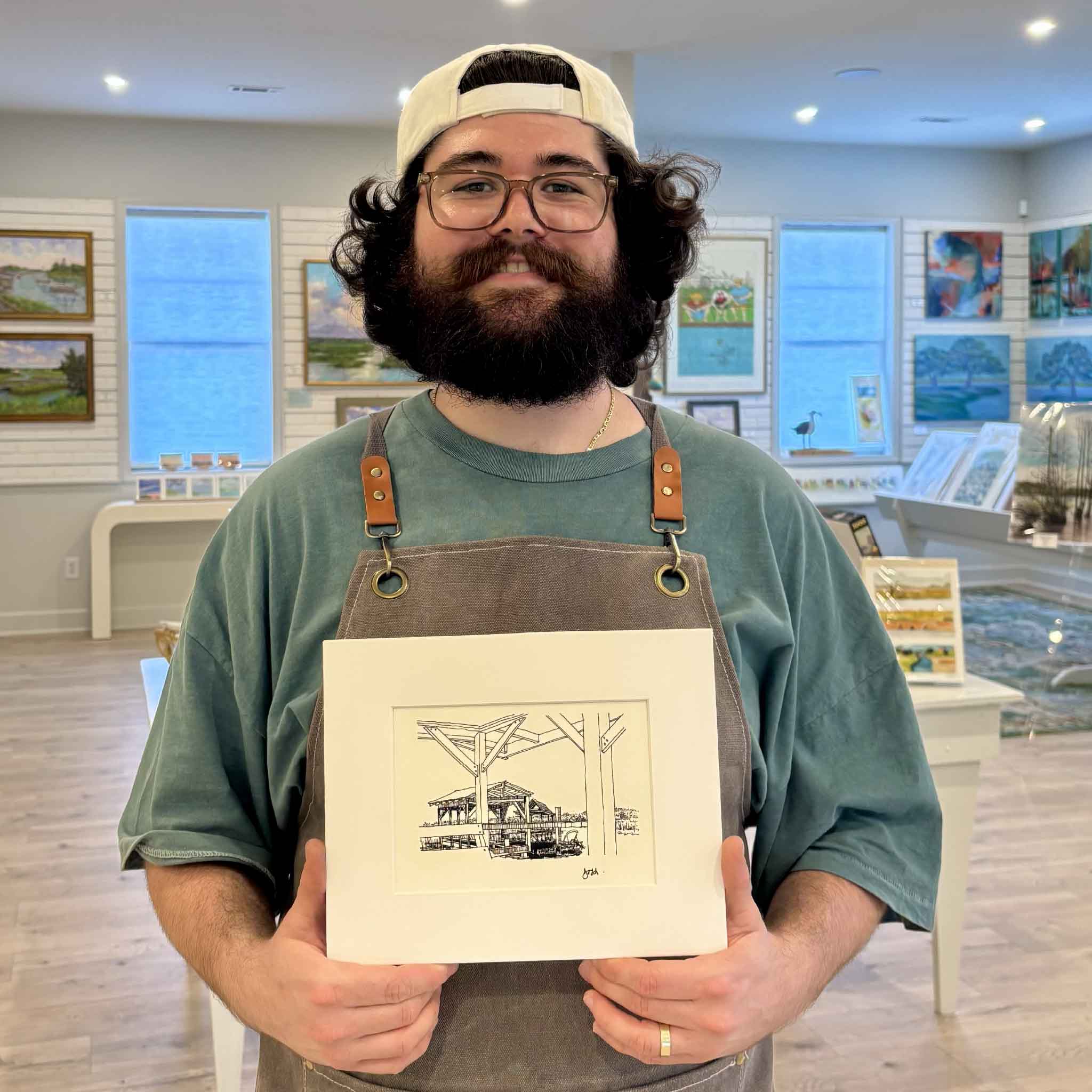 Peyton holds Todd Halamka's Pawleys Island Salt Marsh Looking South pen-and-ink print inside Gray Man Gallery in Pawleys Island, South Carolina. Professionally matted and ready for a standard frame, this detailed coastal print captures one of Pawleys Island's most recognizable salt marsh views, with a dock framing the historic Fish Club pavilion beyond.