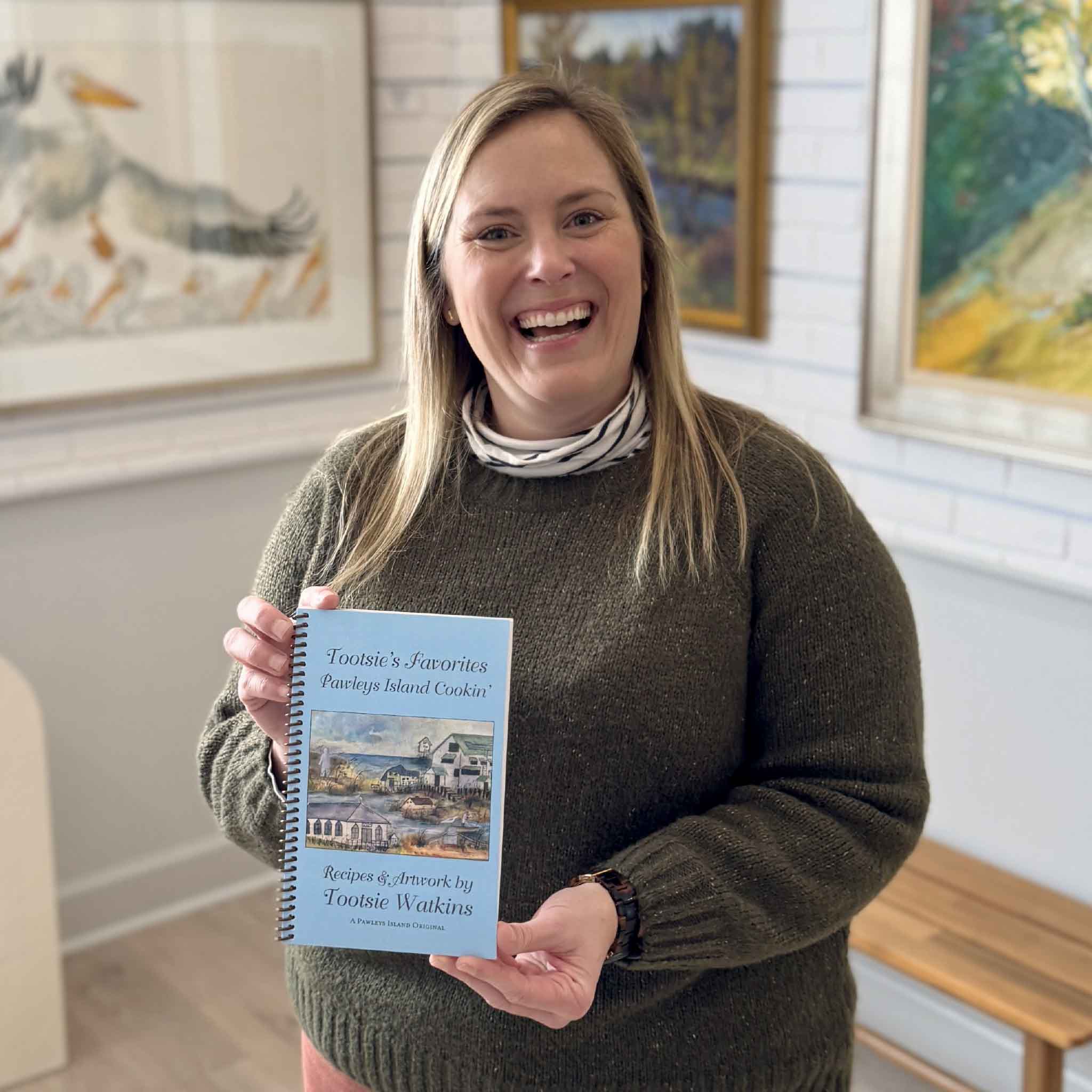 Bordy holding Tootsie’s Favorites: Pawleys Island Cookin’ spiral-bound cookbook by local author and artist Tootsie Watkins at Gray Man Gallery in Pawleys Island, featuring Lowcountry recipes and coastal artwork