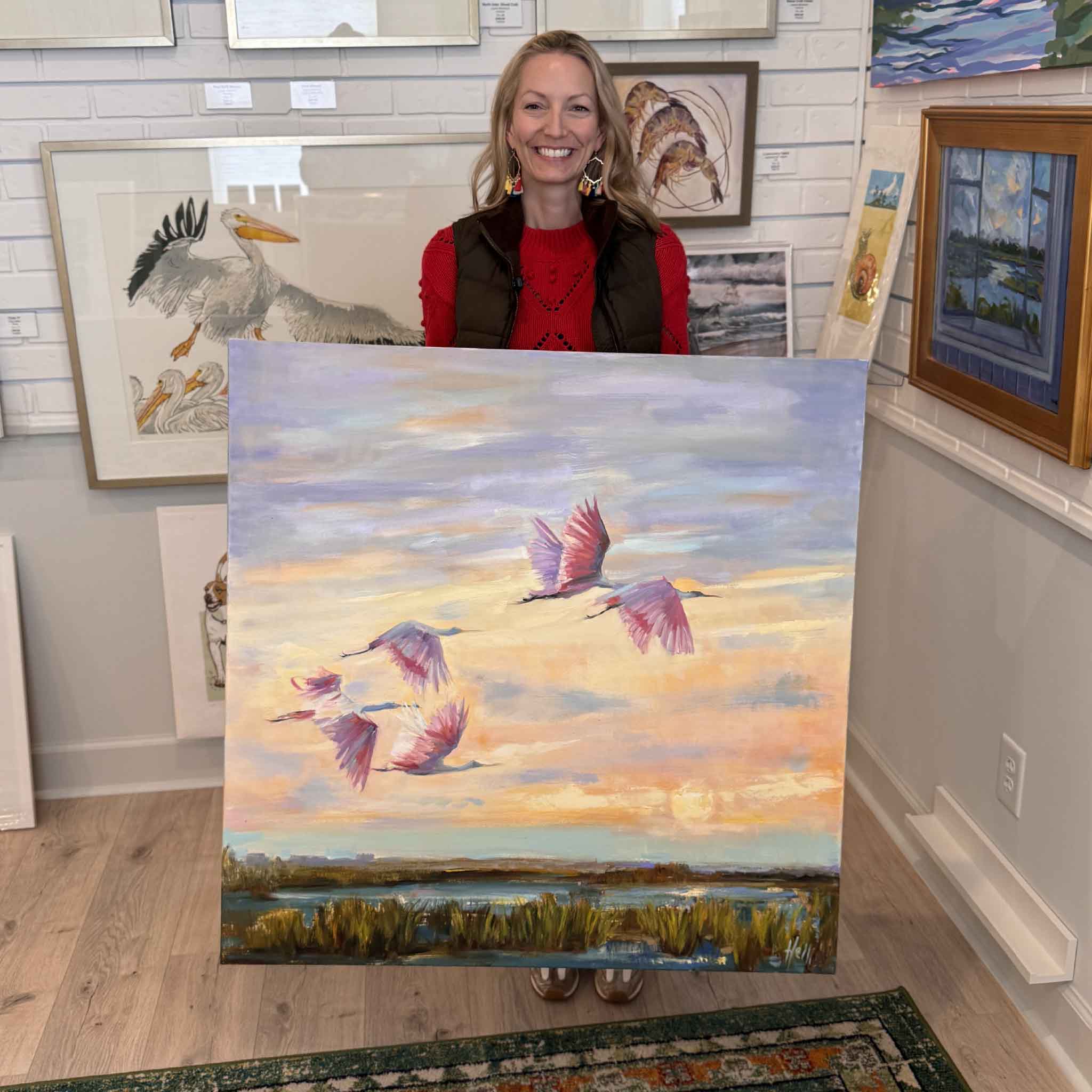 Local artist Jeanne O'Neal holding Twilight Ballet 36x36 oil painting by Helli Luck featuring roseate spoonbills flying over a Lowcountry marsh at sunset
