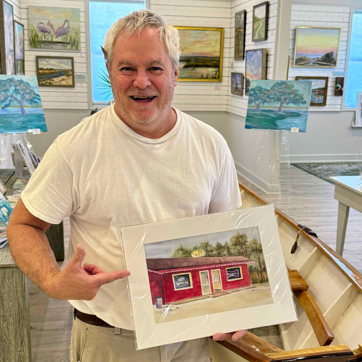 Lee, who remembers visiting the original Club 17 on Highway 17 in Pawleys Island, enthusiastically displaying the 12×16 watercolor print by South Carolina artist Vida Miller. The artwork depicts the legendary red roadside bar with its familiar vintage beer signs, preserving a colorful piece of local history. Photographed inside Gray Man Gallery in Pawleys Island, South Carolina.