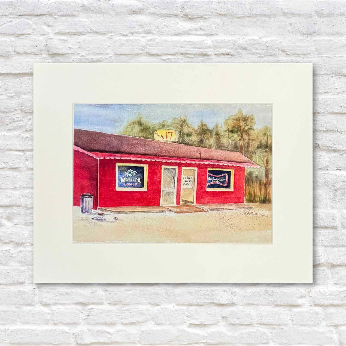 Club 17 fine art watercolor print by local artist and Gray Man Gallery founder Vida Miller, depicting the iconic red Club 17 roadside landmark on Highway 17 in Pawleys Island, South Carolina, with vintage beer signs, a red roof, and wooded backdrop. Matted print photographed against a white brick wall at Gray Man Gallery.