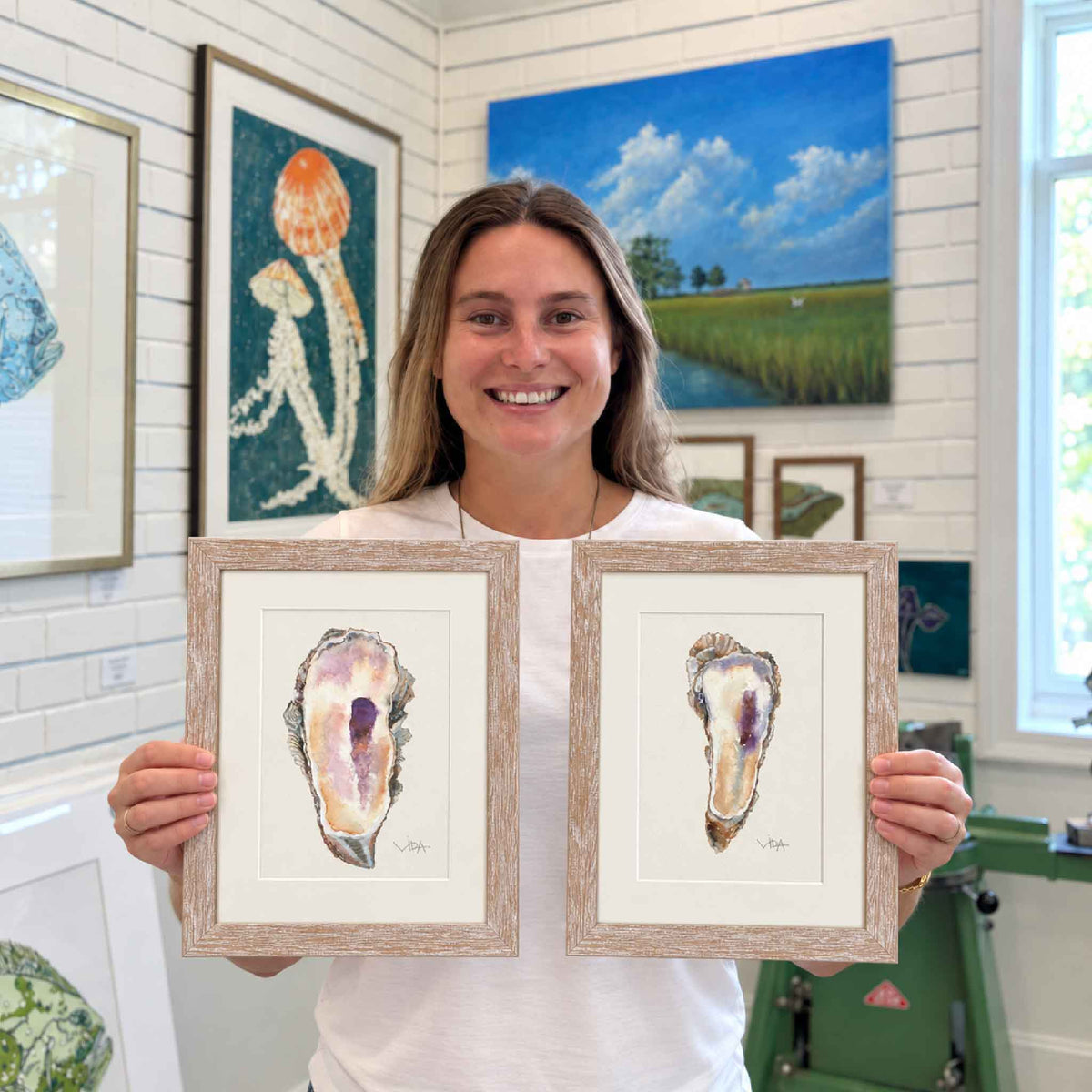Kyra holds Pawleys Oyster I and Pawleys Oyster II, two one-of-a-kind original watercolor paintings of Lowcountry oyster shells by Pawleys Island artist Vida Miller, each professionally matted and framed in a softly weathered light wood frame, photographed inside Gray Man Gallery in Pawleys Island, South Carolina