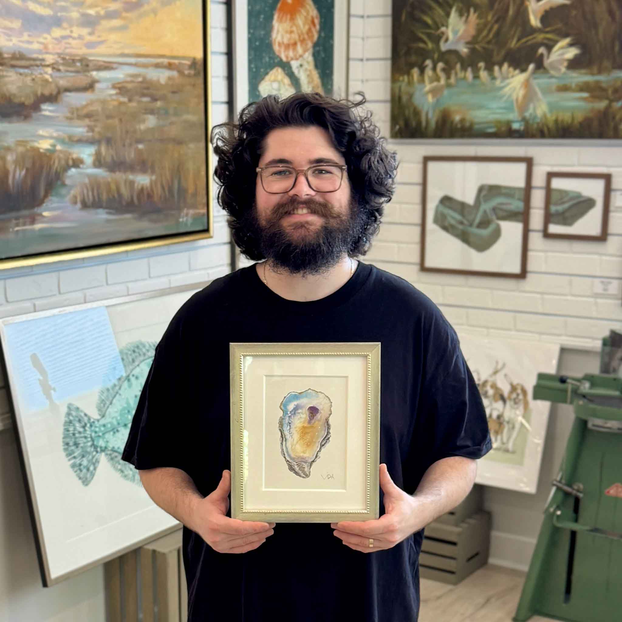 Peyton holding Pawleys Oyster V, a one-of-a-kind original watercolor oyster painting by local artist Vida Miller, professionally matted and custom framed at Gray Man Gallery in Pawleys Island, South Carolina, with original Lowcountry artwork displayed throughout the gallery behind him.