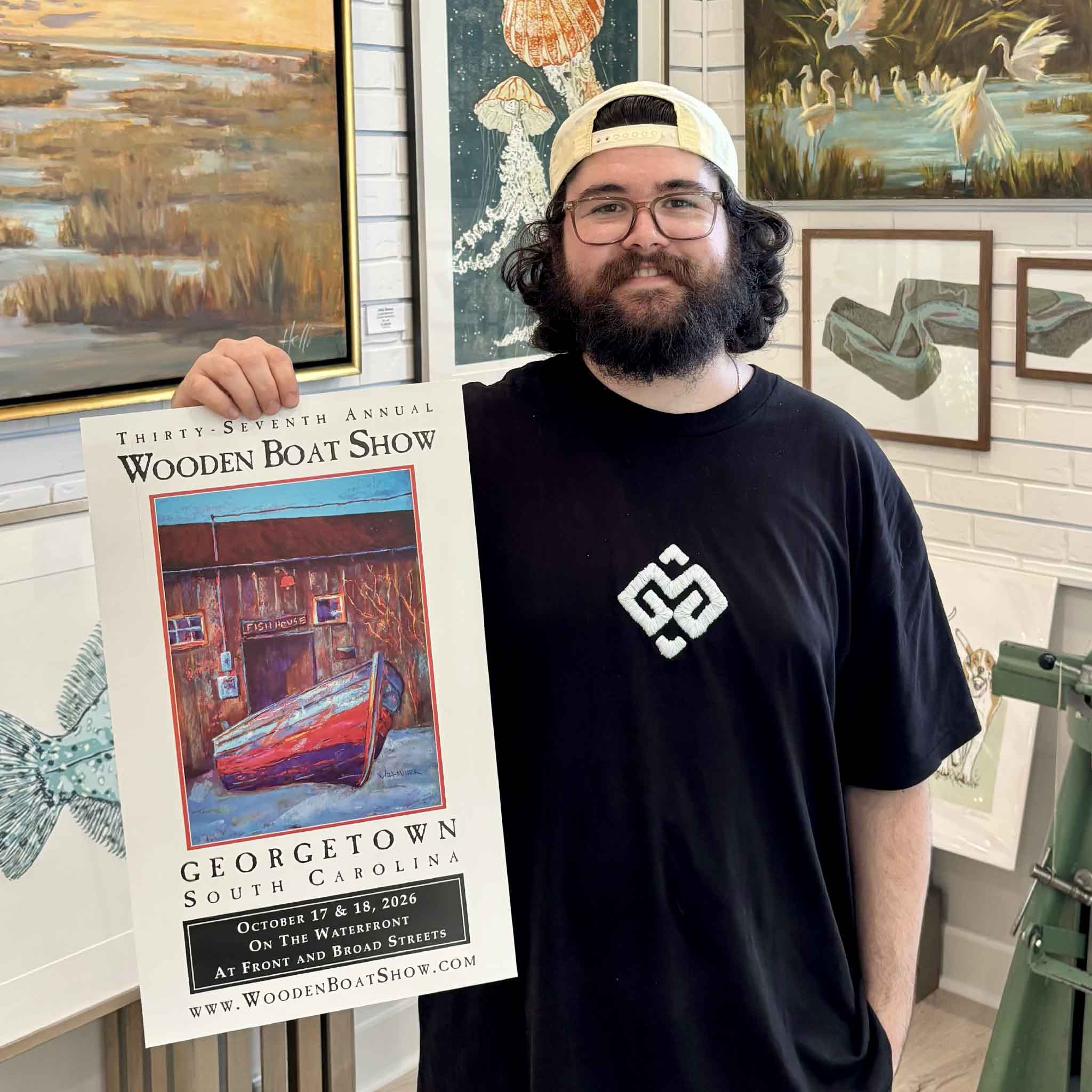 Peyton holds the official signed 2026 Georgetown Wooden Boat Show poster by South Carolina artist Vida Miller inside Gray Man Gallery in Pawleys Island. The poster features Miller’s vibrant painting of the historic Cathou Fish House and wooden boat on the Georgetown, South Carolina waterfront.