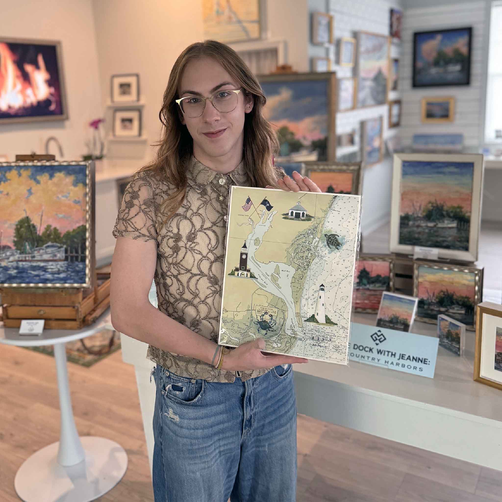 Team member holding Vida Miller's illustrated Winyah Bay map inside Gray Man Gallery in Pawleys Island, South Carolina. The artwork features Georgetown, Pawleys Island, Winyah Bay, nautical chart details, the Georgetown Lighthouse, Pawleys Island Chapel, wildlife, and coastal landmarks.