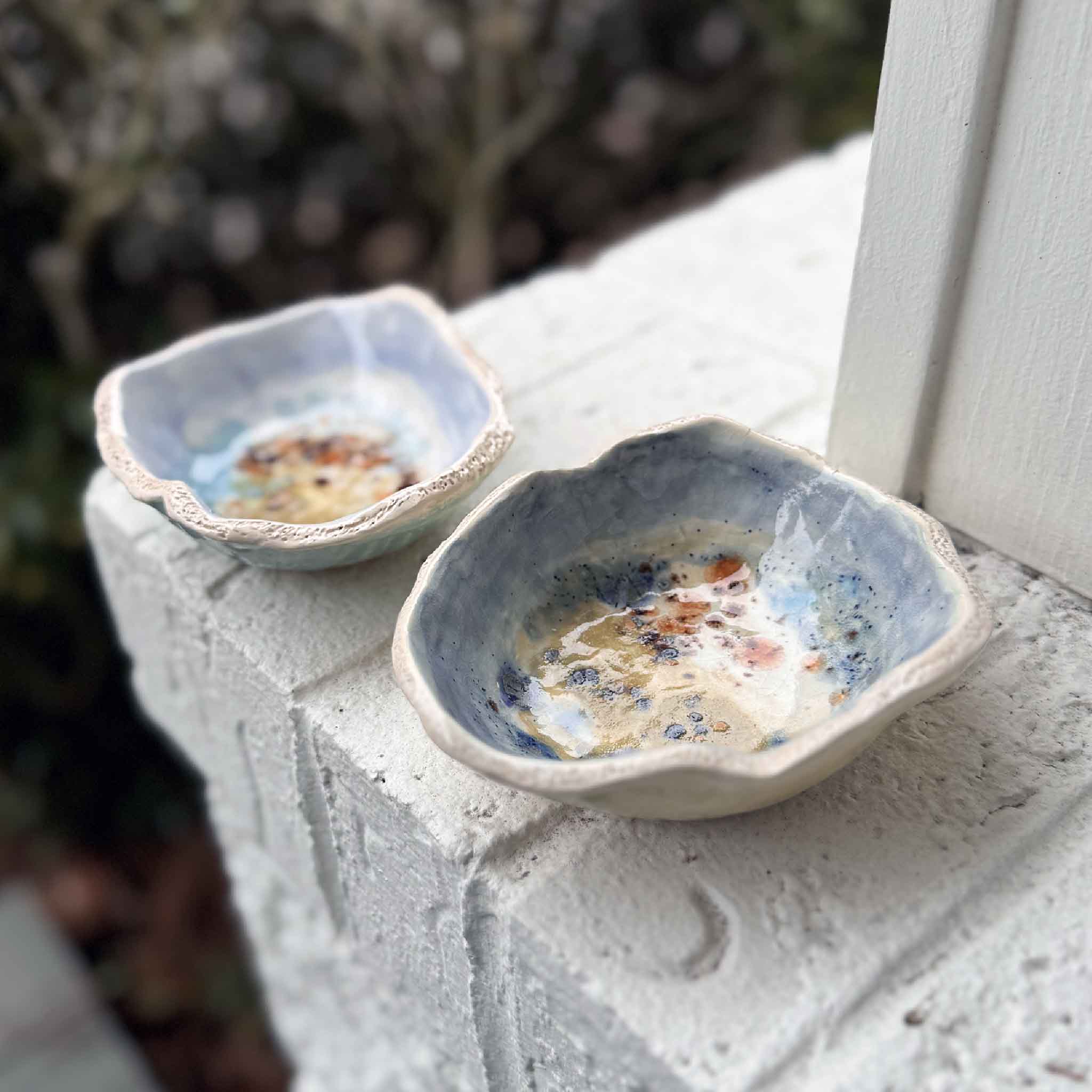 Two small light blue handcrafted pottery bowls by Vida Miller, styled together on a white brick ledge at Gray Man Gallery near coastal greenery.