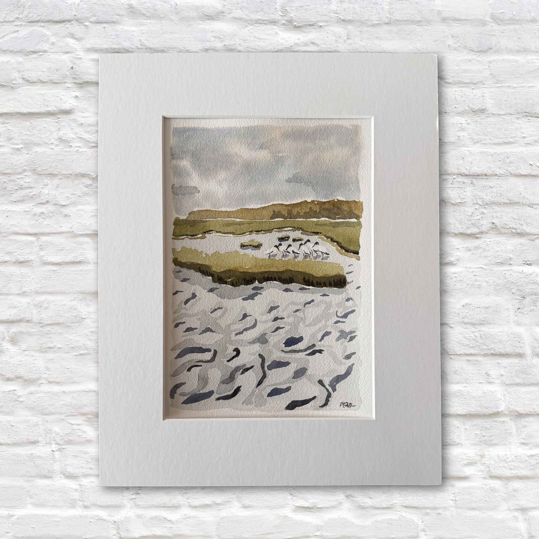 Original watercolor titled Woodstorks at the Horseshoe by local artist Maggie Pelton, depicting a group of wood storks gathered along a winding Lowcountry marsh waterway under soft coastal skies. The artwork measures 7x10 inches and is professionally matted to fit a standard 11x14 frame, shown matted against a white brick backdrop at Gray Man Gallery in Pawleys Island, South Carolina.