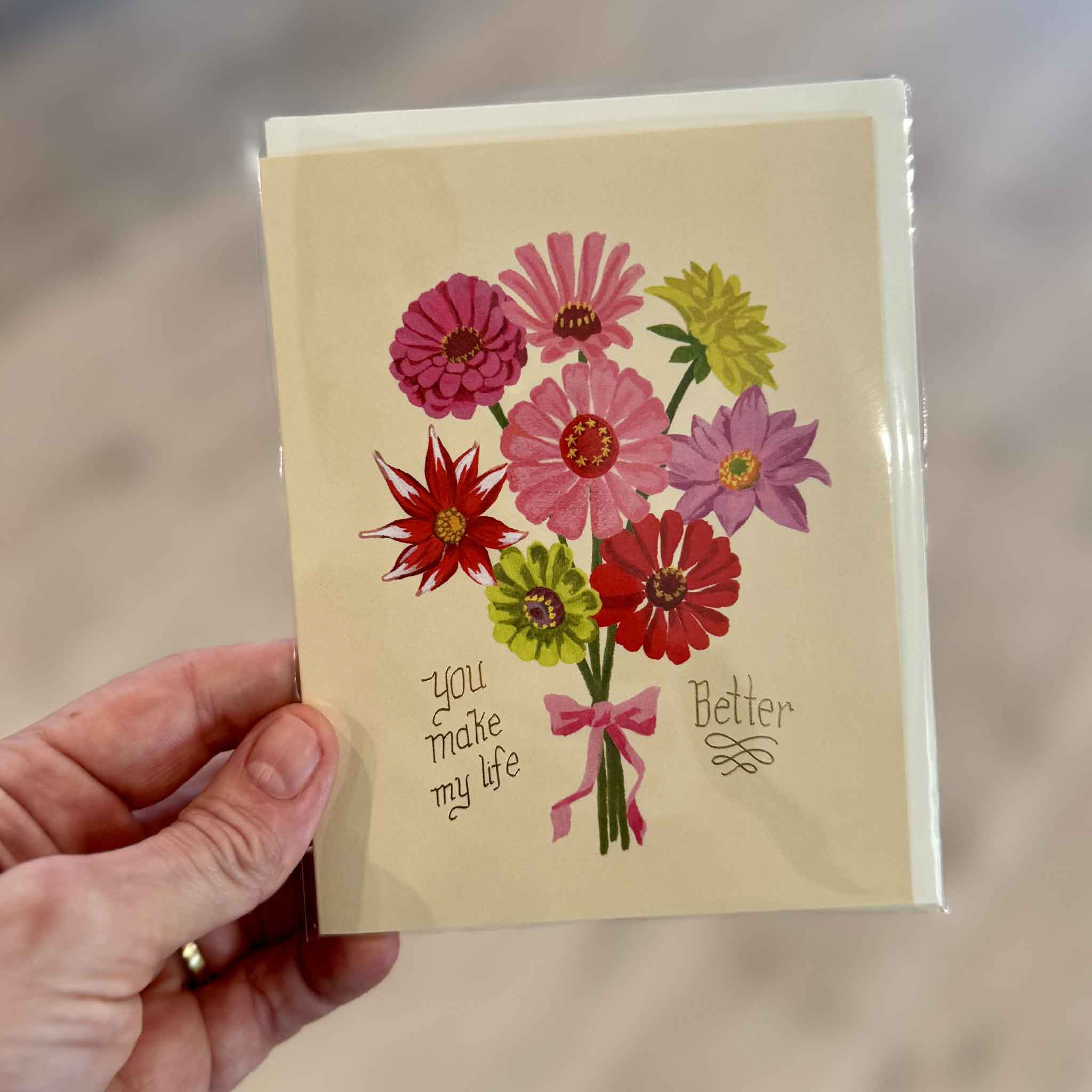 Hand holding the You Make My Life Better greeting card featuring a bright bouquet of pink, red, purple and green flowers with a pink ribbon and metallic gold foil message, a sweet love, friendship or just-because card available at Gray Man Gallery in Pawleys Island, South Carolina.