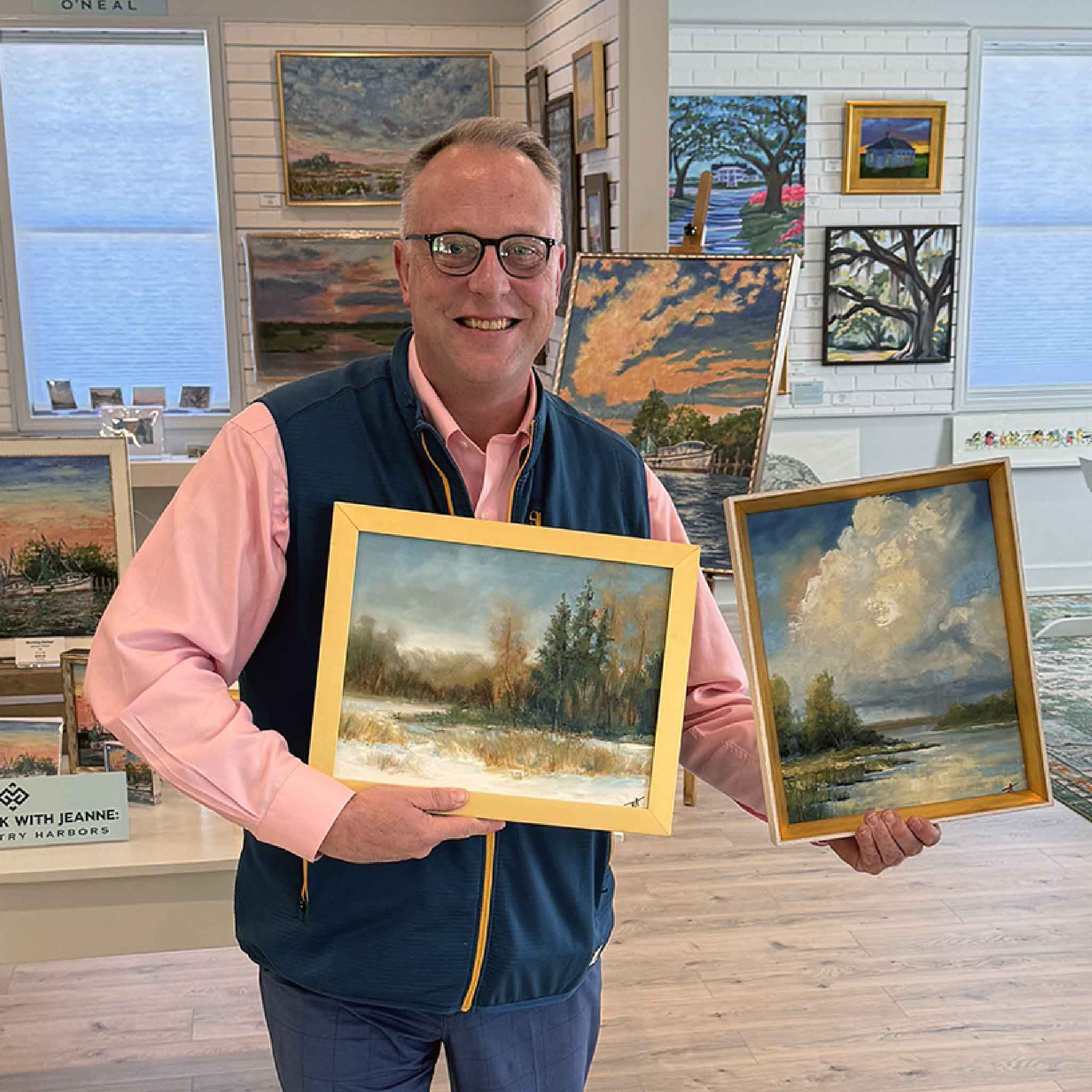 Artist and meteorologist Jamie Arnold standing inside Gray Man Gallery, holding two framed Lowcountry landscape oil paintings from his coastal collection.