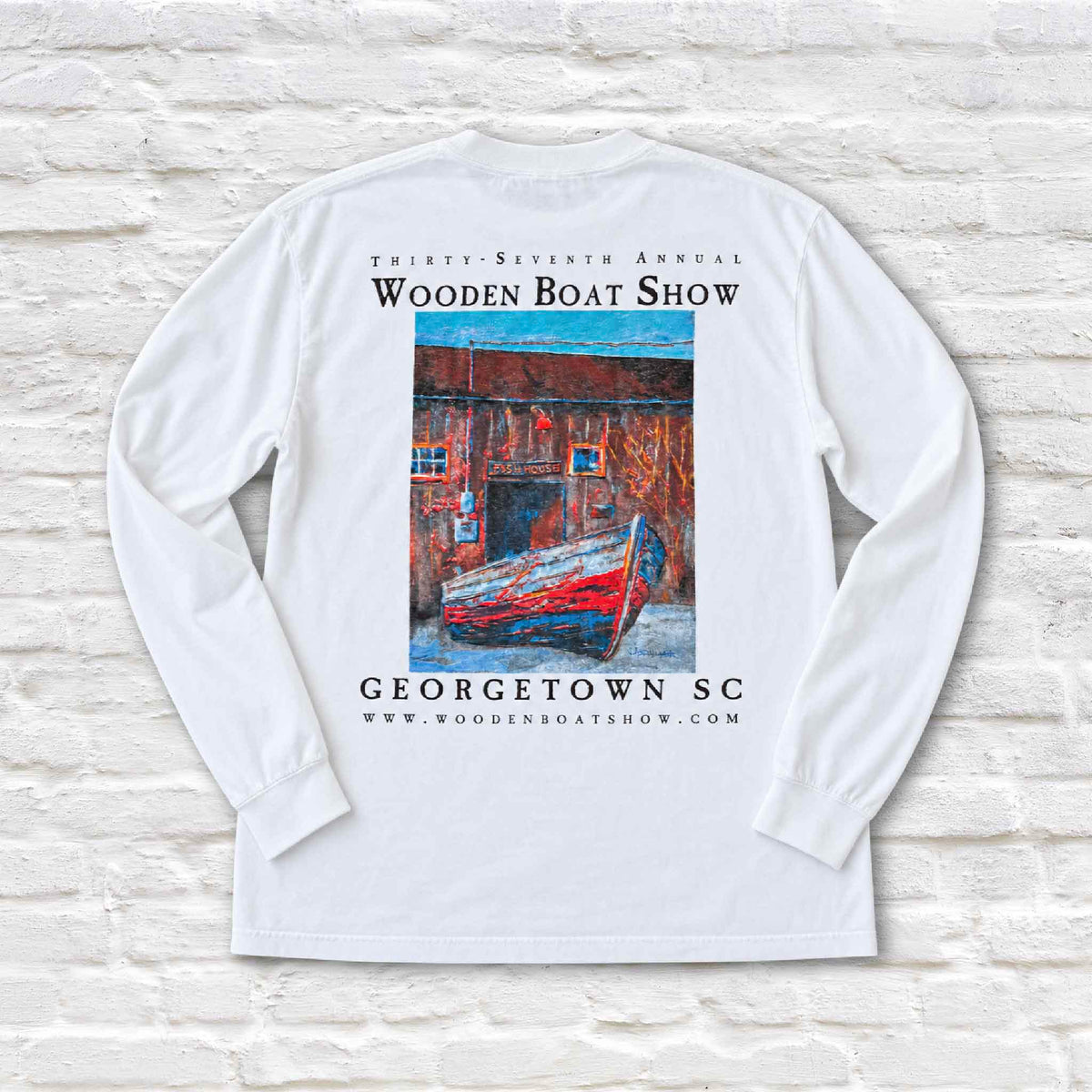 Official 2026 Georgetown Wooden Boat Show white long sleeve Comfort Colors T-shirt photographed flat against Gray Man Gallery's white brick background. The back features a full-color reproduction of local South Carolina artist Vida Miller's painting of the historic Georgetown Fish House and wooden boat, with lettering celebrating the 37th Annual Wooden Boat Show in Georgetown, South Carolina.
