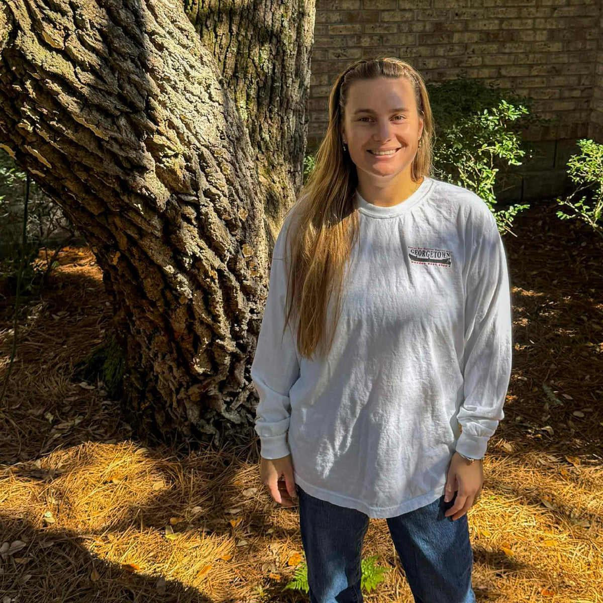 Front view of the official 2026 Georgetown Wooden Boat Show white heavyweight Comfort Colors long sleeve cotton T-shirt modeled outdoors at Gray Man Gallery in Pawleys Island, South Carolina. Features a small Georgetown Wooden Boat Show logo on the left chest, a classic crewneck, and relaxed long sleeve styling.