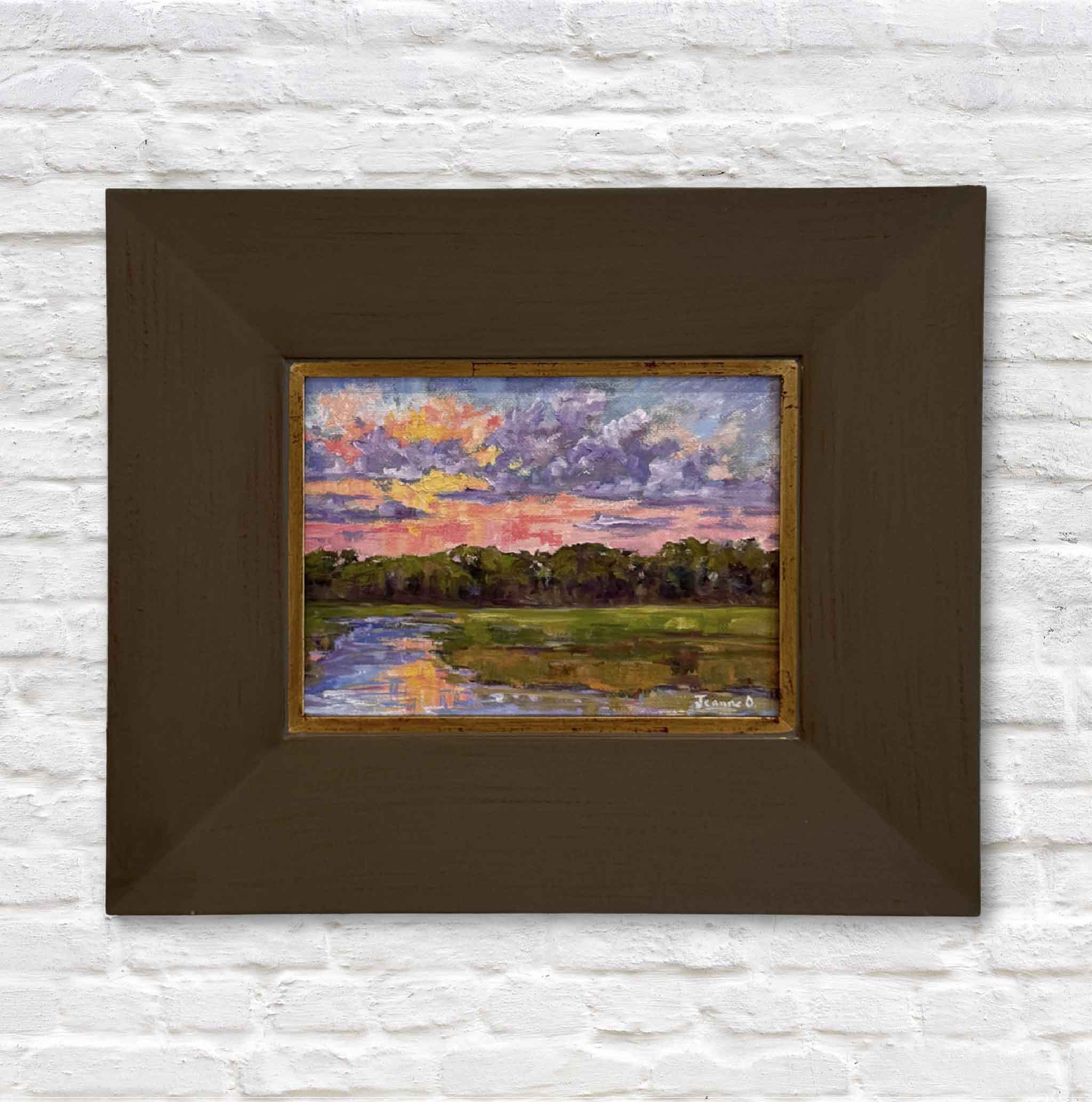 Framed painting of a landscape with a colorful sky on a white brick wall.