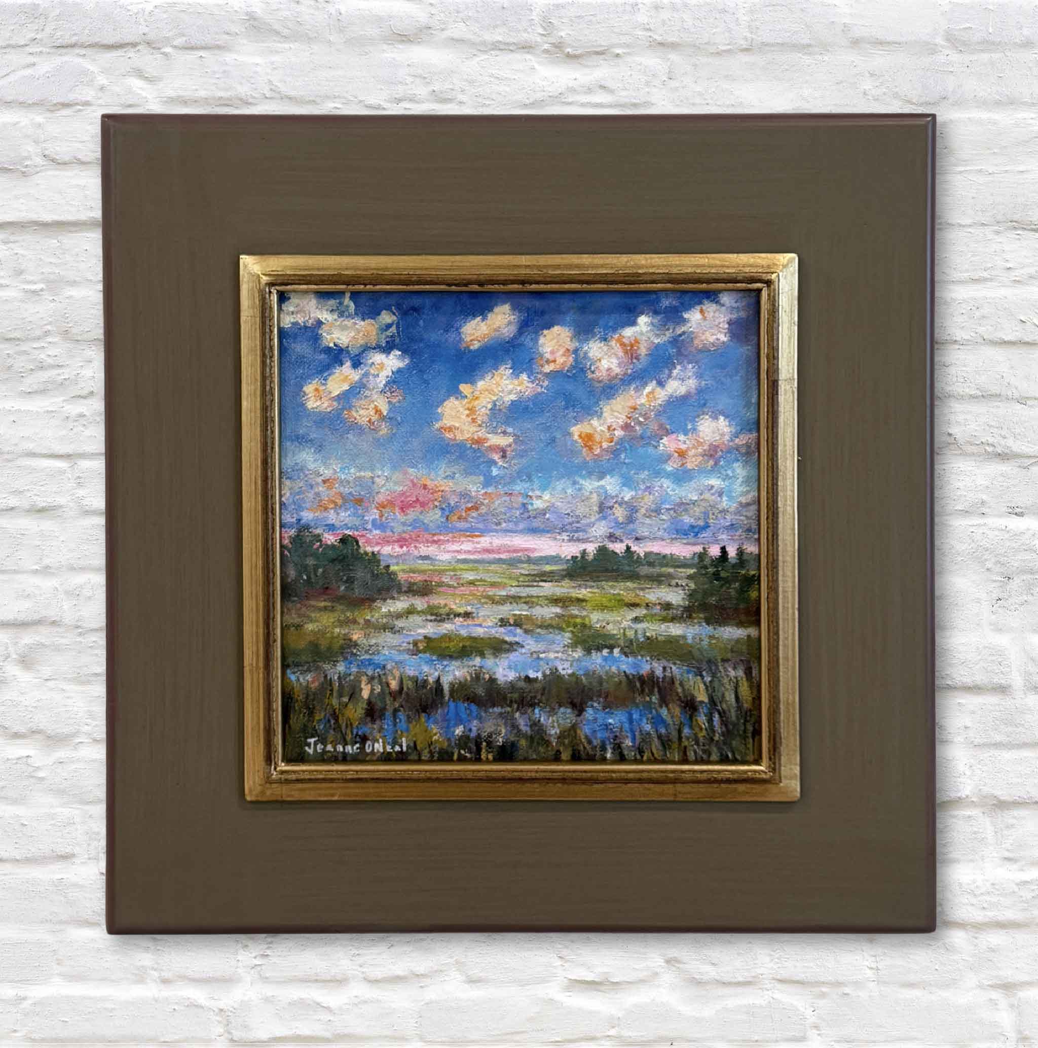 Framed painting of a landscape with a blue sky and clouds on a white brick wall.