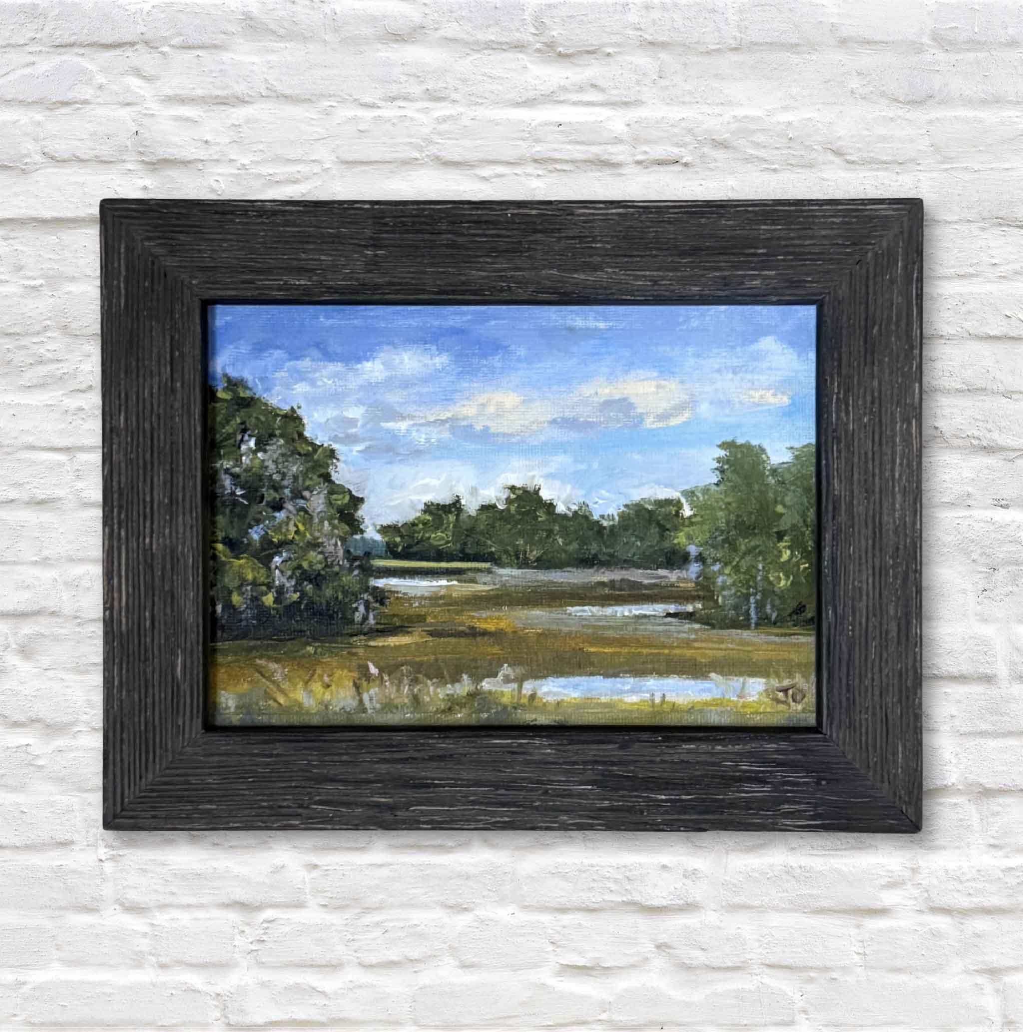 Framed painting of a landscape with trees and sky on a white brick wall.