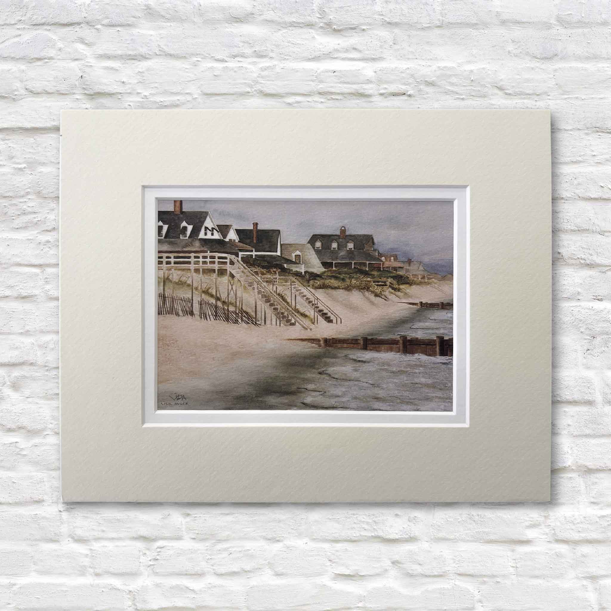 matted artwork of a coastal beach scene with houses and a beach on a white brick wall.