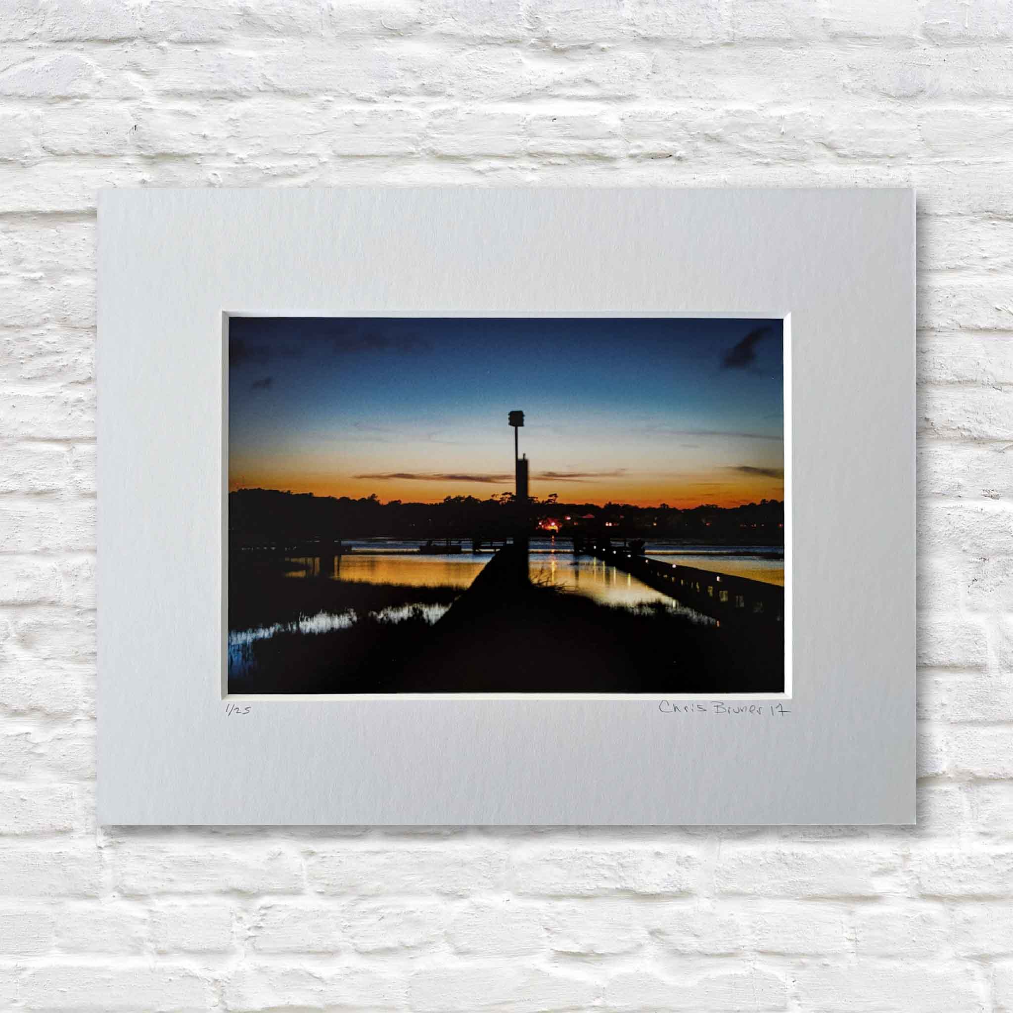 matted photograph of a sunset over a dock with a birdhouse, mounted on a white brick wall.