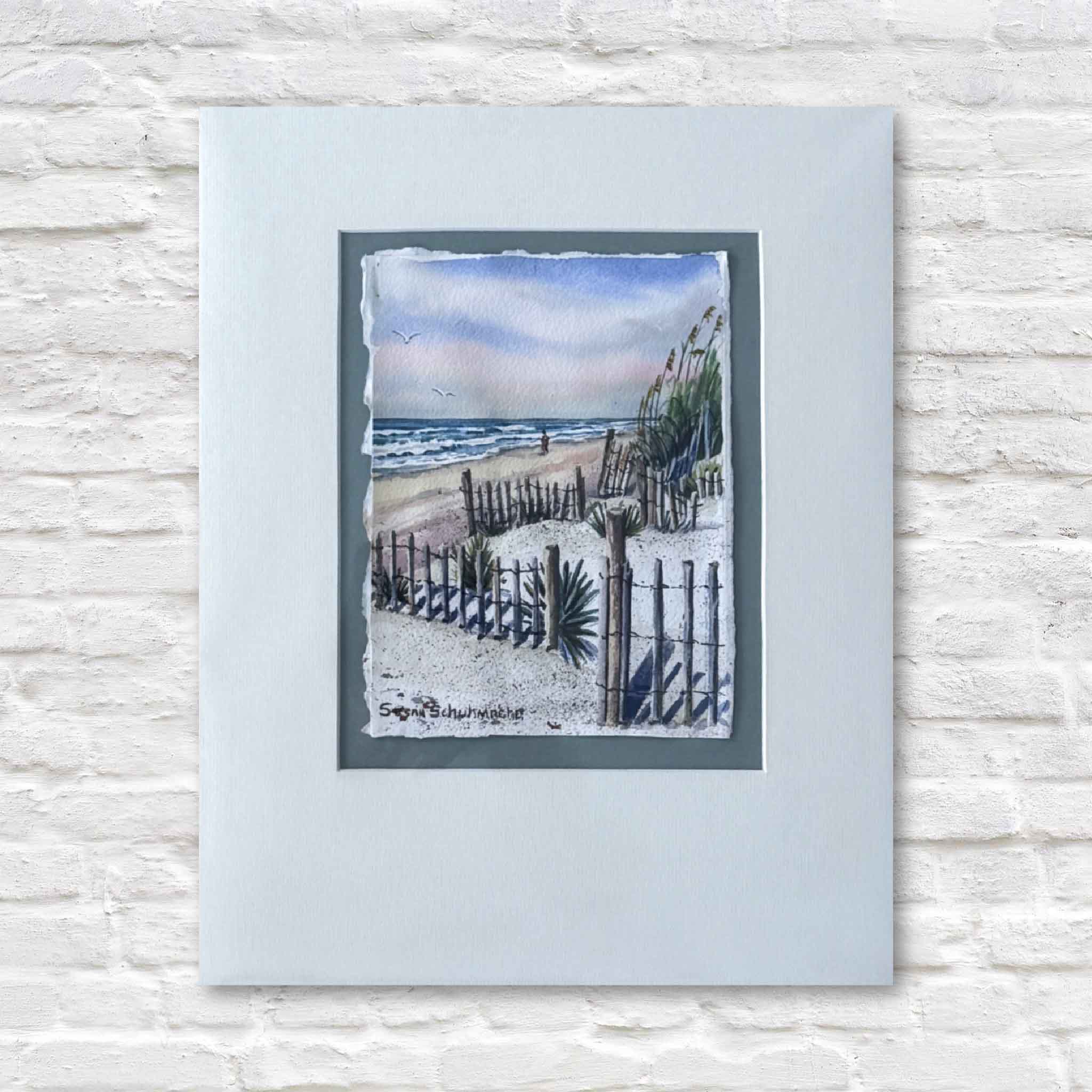 matted artwork of a beach scene with a fence and ocean view on a textured white surface.