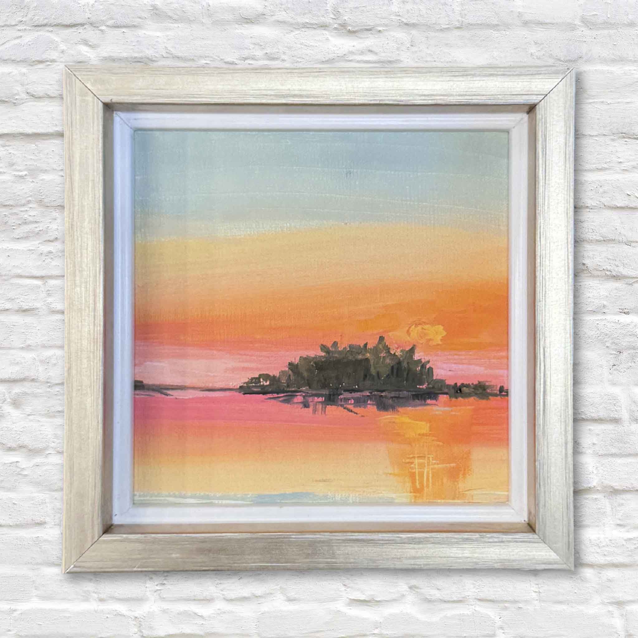 Framed artwork of a sunset over water with an island, on a textured white wall.