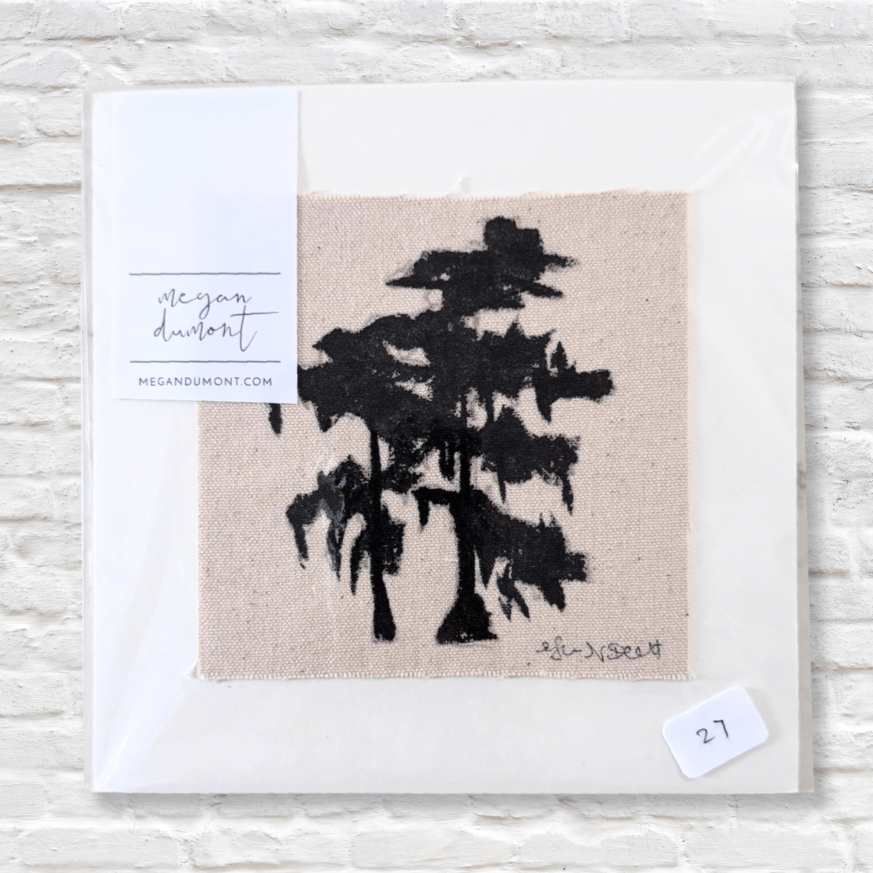 Art print with black tree silhouette on beige fabric, signed 'Megan Dumont', against a light wood background.