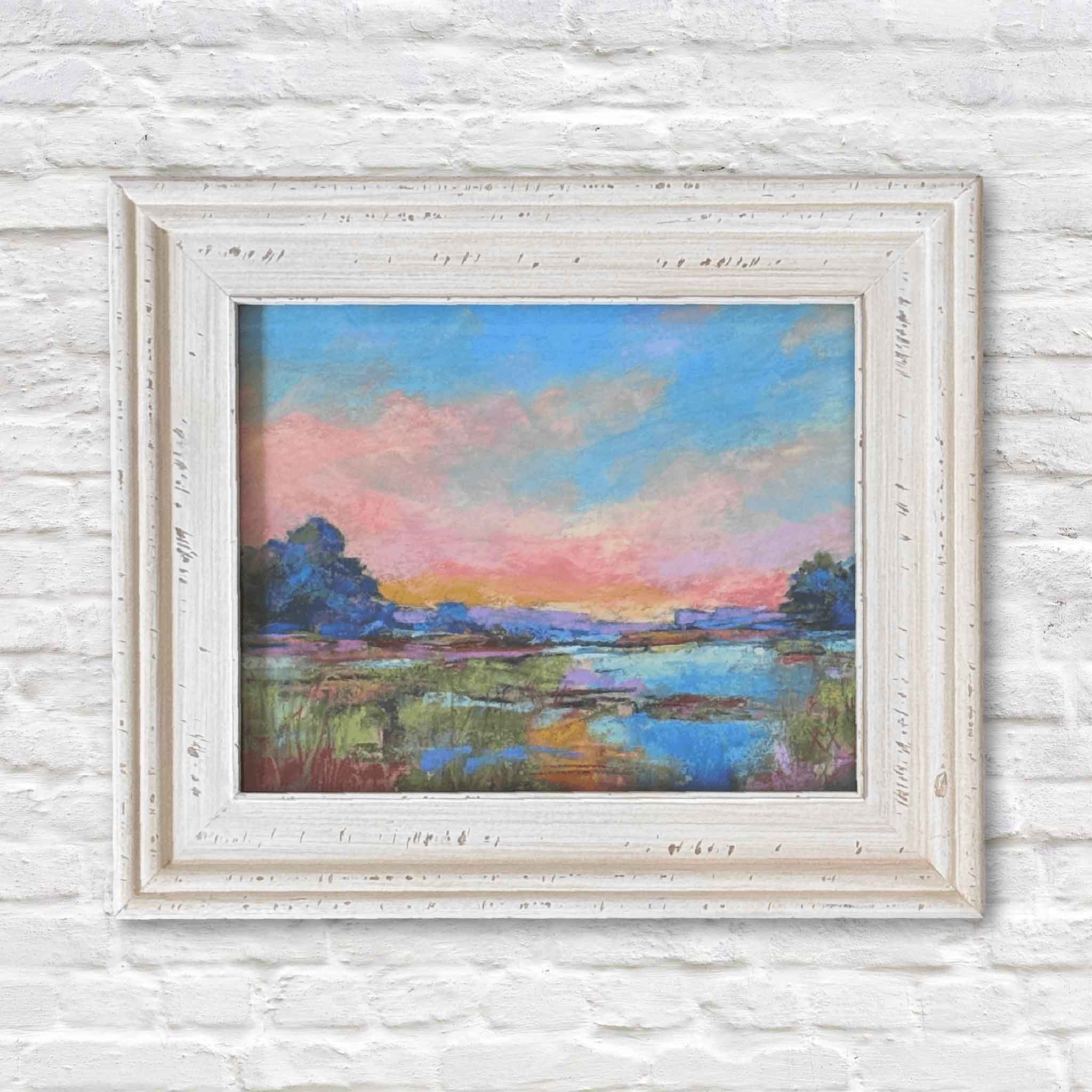 Framed painting of a landscape with a sunset over water on a white textured wall.