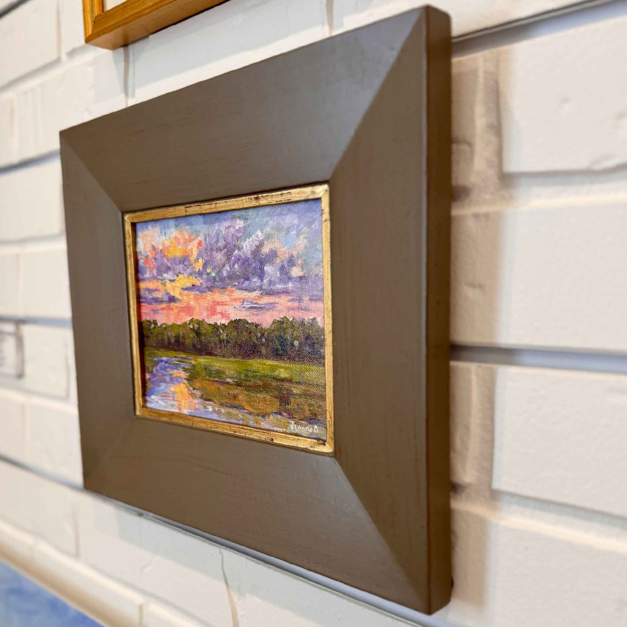 Framed painting of a sunset over water on a white brick wall.