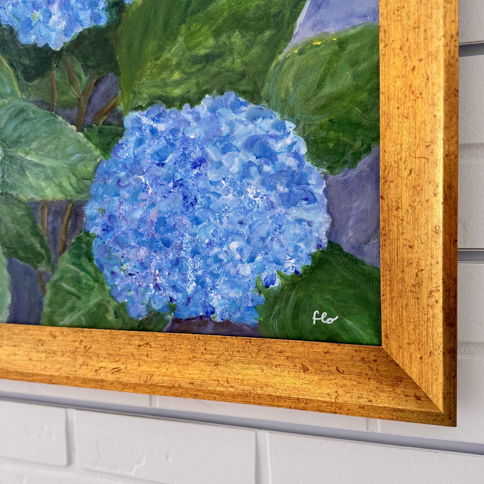 Framed painting of blue hydrangeas with artist's signature on a white brick wall