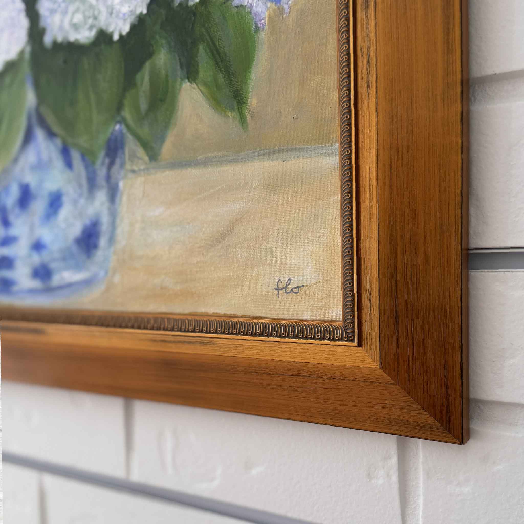 Framed painting of flowers with artist's signature on a white brick wall