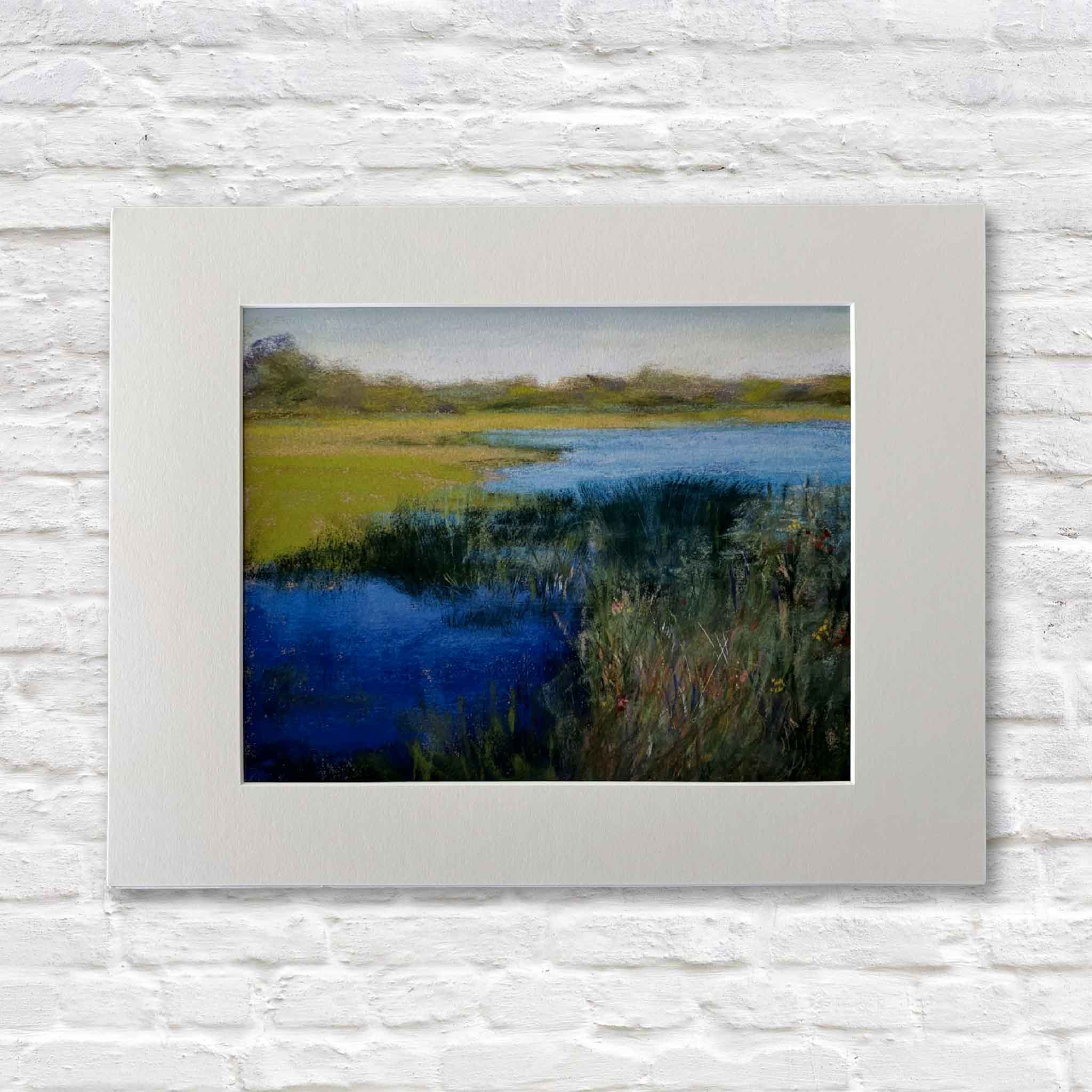 matted painting of a landscape with blue water and green fields on a white brick wall.