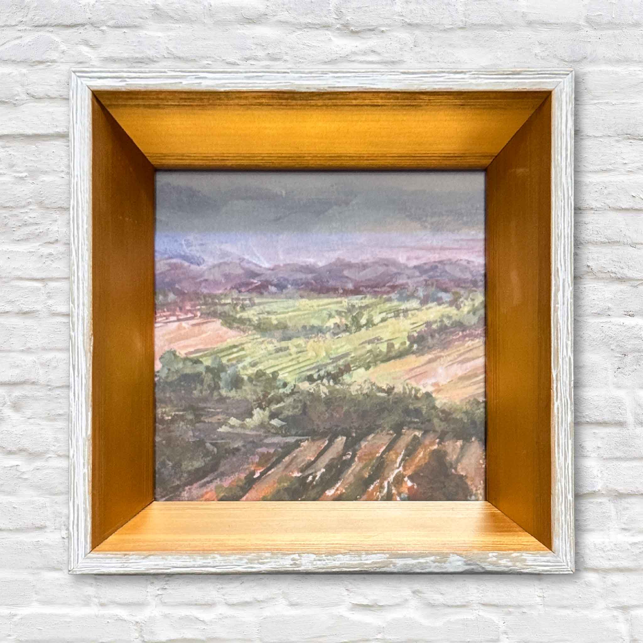 Framed painting of a landscape with a wooden frame on a textured white wall.