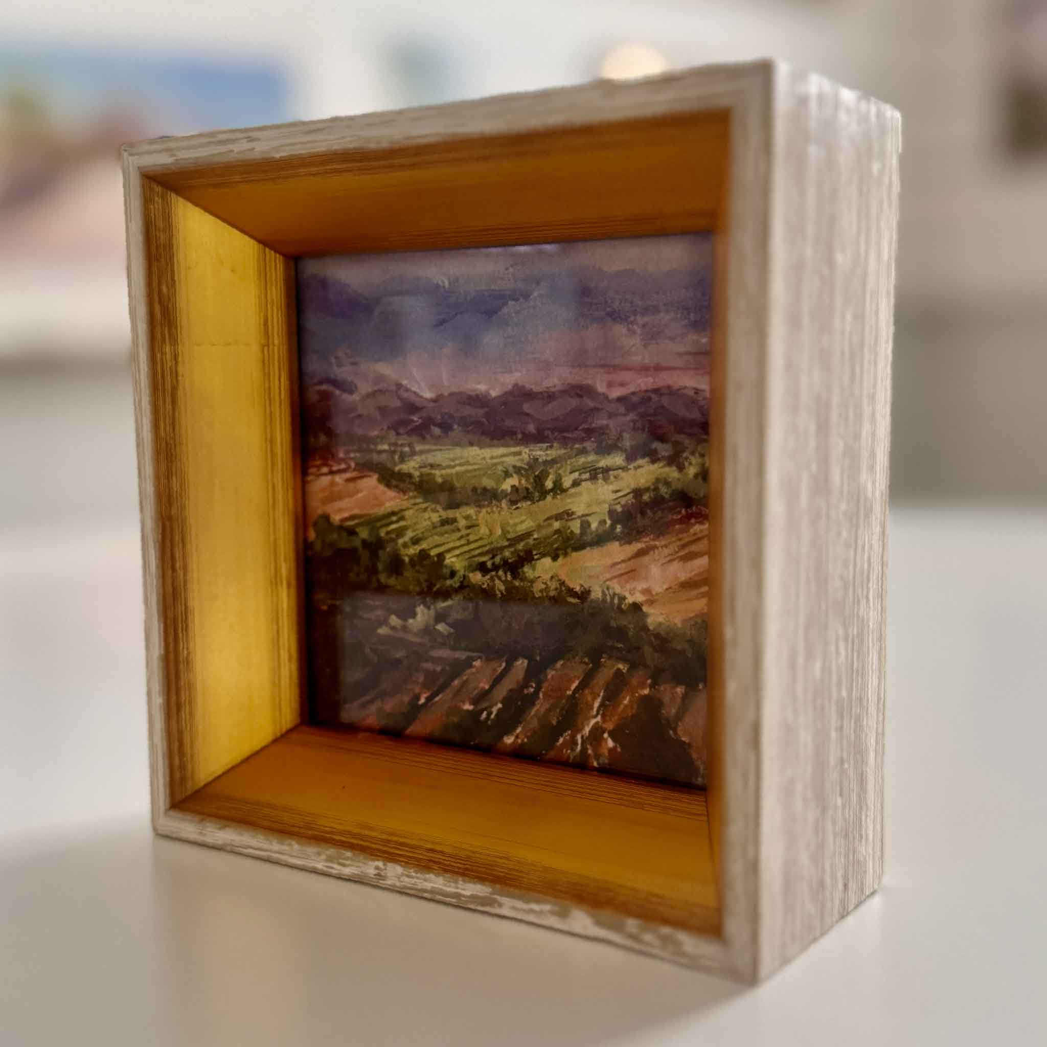 Small landscape painting inside a wooden frame on a white surface