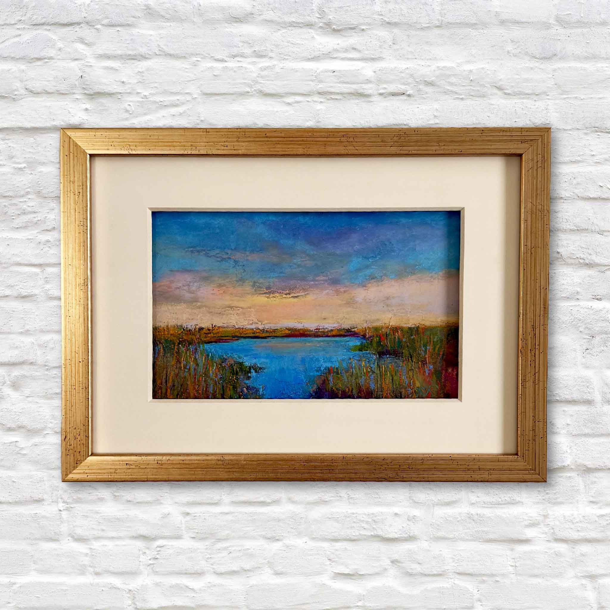 Framed pastel marsh landscape painting by Joanne Willoughby, featuring a glowing Lowcountry sunset over blue tidal waters and golden grasses, matted in cream with a gold frame