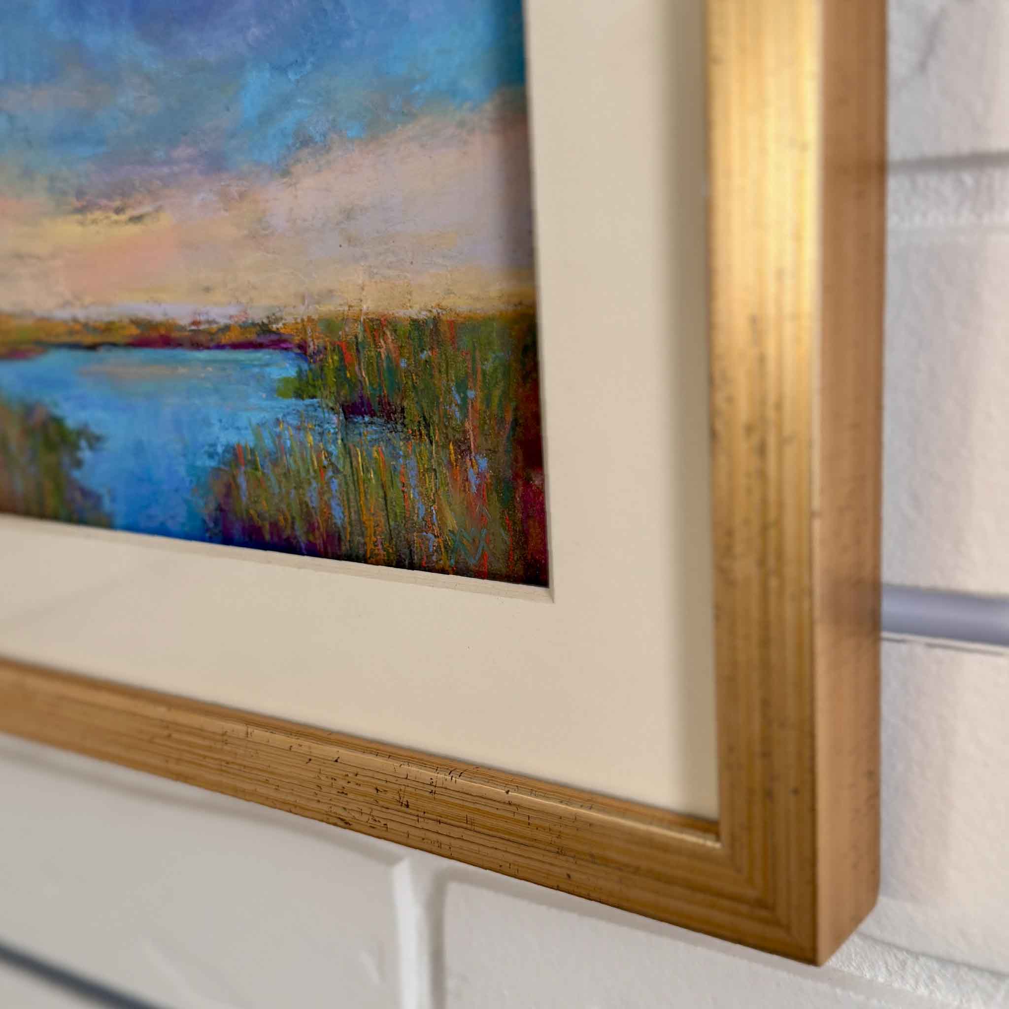 Framed pastel marsh landscape painting by Joanne Willoughby, featuring a glowing Lowcountry sunset over blue tidal waters and golden grasses, matted in cream with a gold frame
