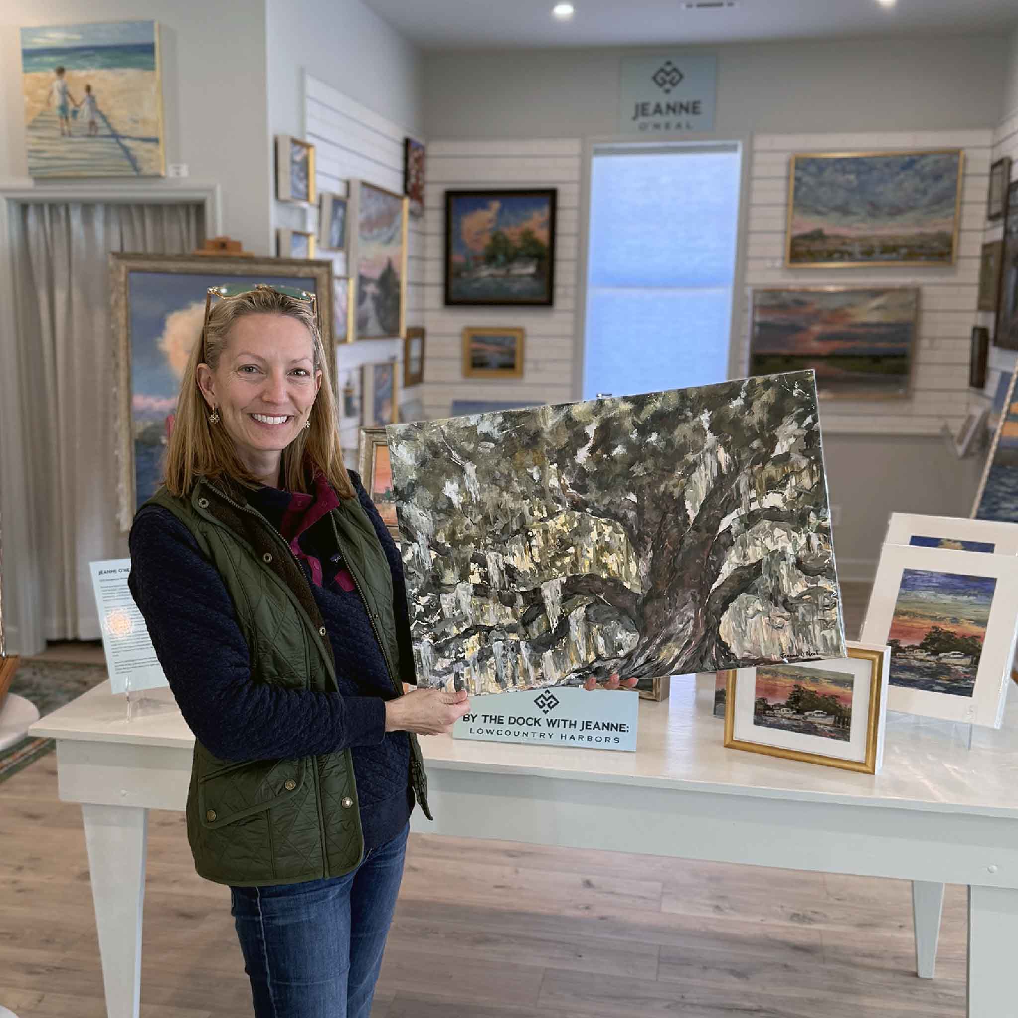 Artist Jeanne O’Neal holding her 24x16 art print Deep Roots inside Gray Man Gallery, featuring a textured painting of a majestic live oak tree bathed in soft Lowcountry light.