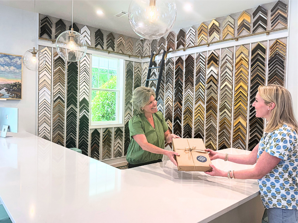 A smiling Gray Man Gallery customer receives a neatly wrapped, custom-framed artwork across the counter from a gallery team member, with a backdrop of sample frame mouldings.
