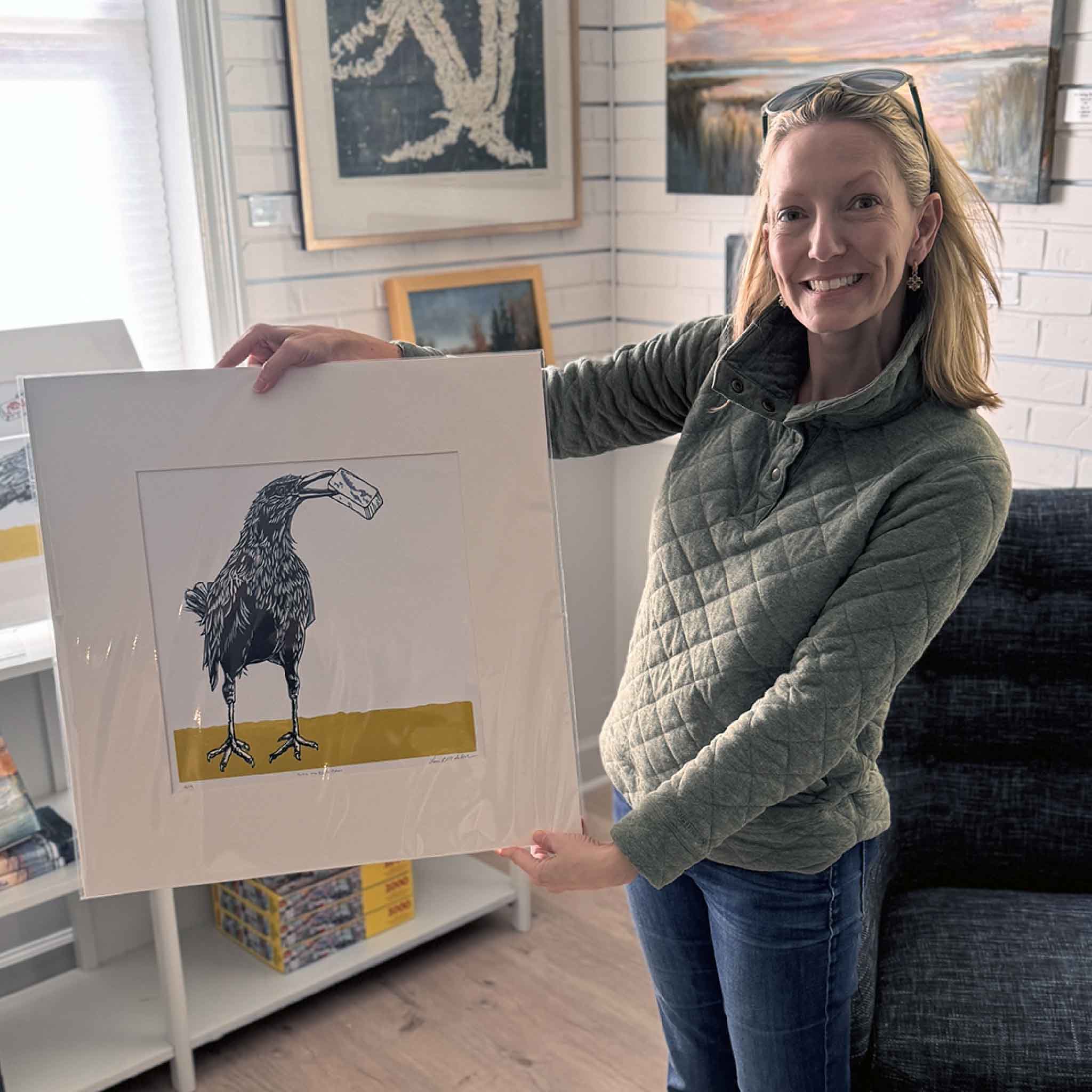 Local artist Jeanne O'Neal standing inside Gray Man Gallery, smiling while holding a large matted black-and-white bird linocut (by Laurie McIntosh) with a mustard-yellow accent. Colorful coastal and abstract paintings hang behind her.