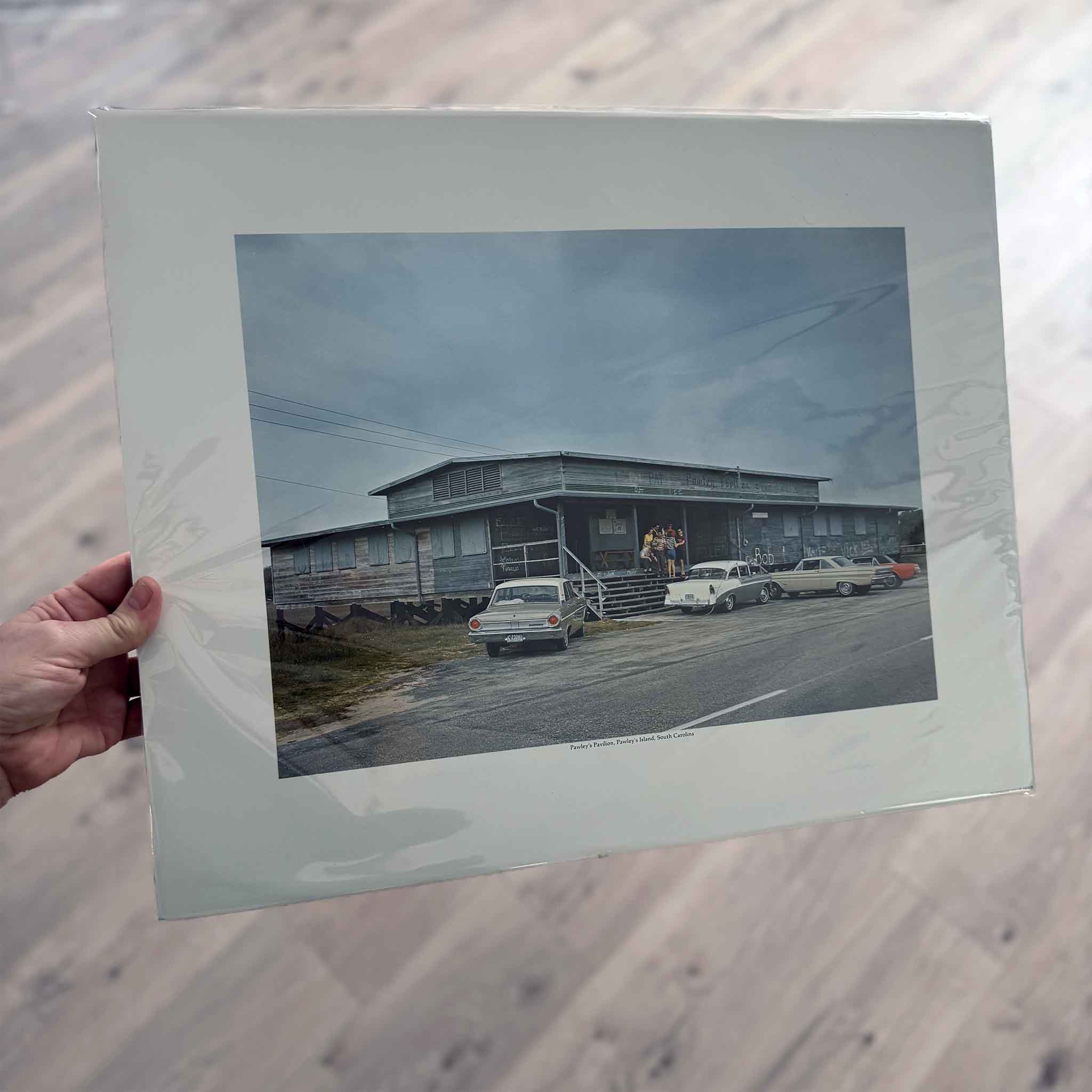 Vintage photograph of Pawleys Pavilion on Pawleys Island, South Carolina, showing classic 1960s cars parked outside a weathered beach building beneath a cloudy coastal sky.