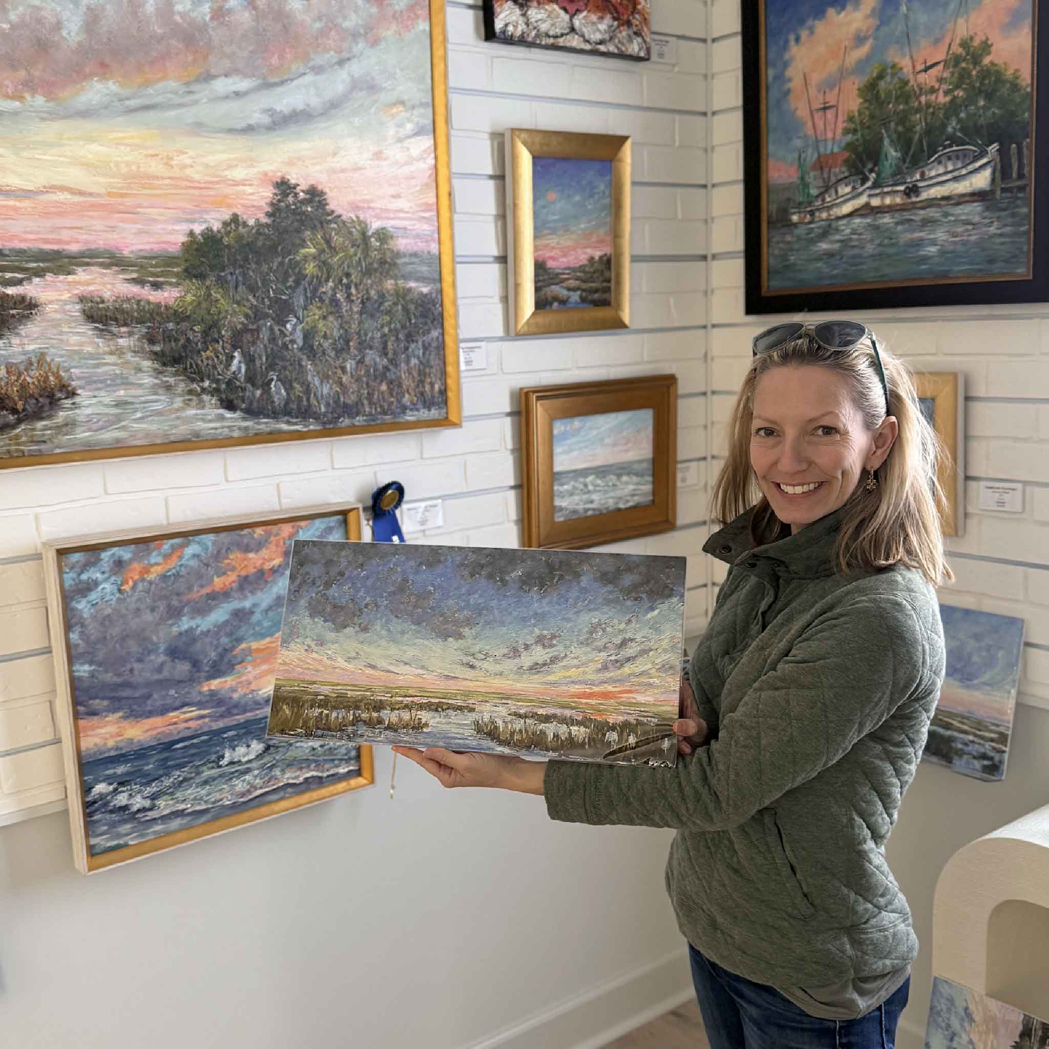 Local Artist Jeanne O'Neal in Gray Man Art Gallery holding a coastal landscape print with a dramatic sky and marsh scene; framed Lowcountry paintings hang on the wall behind her.
