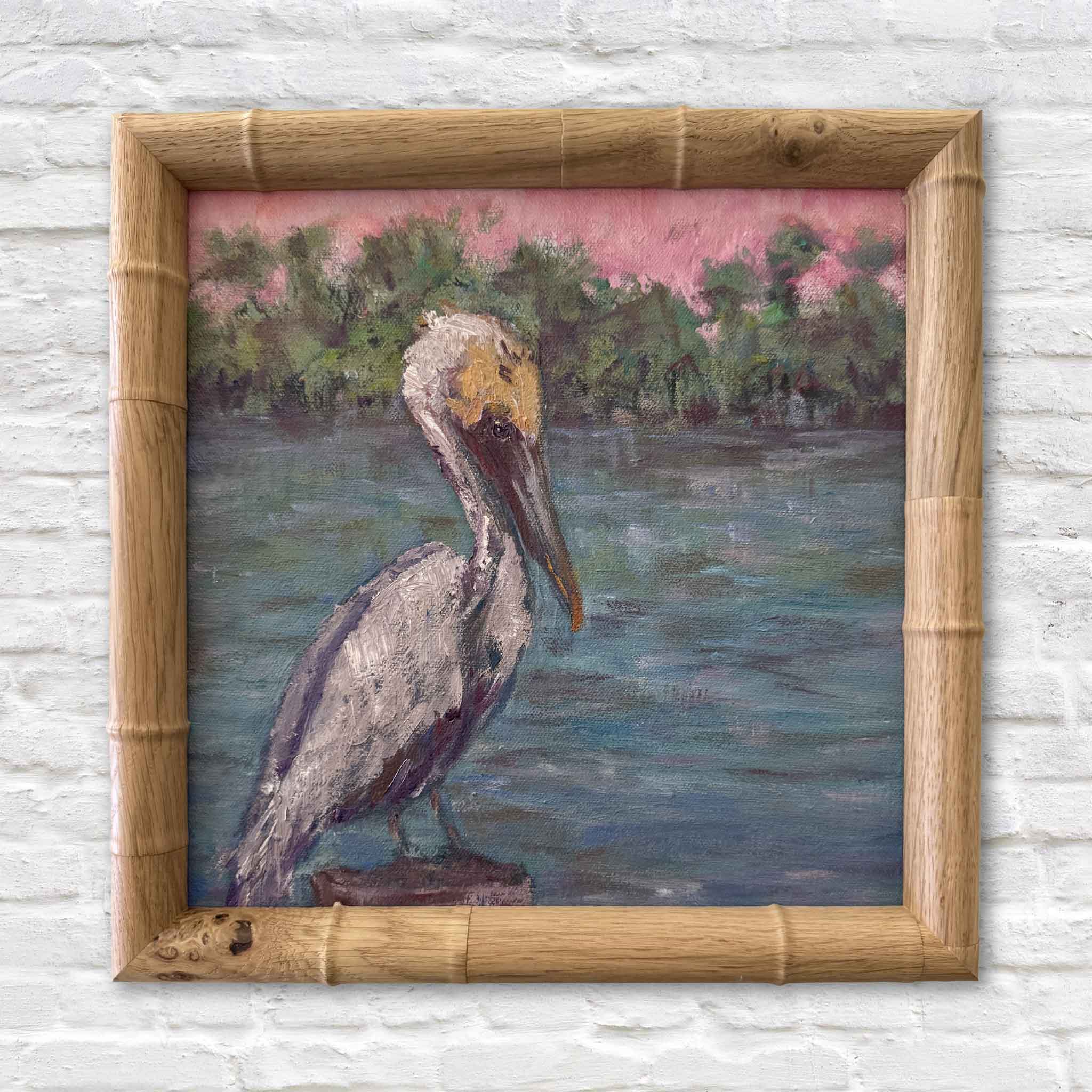 A Moment Alone by Jeanne O'Neal, an original oil painting of a pelican perched beside calm Lowcountry waters beneath a pink evening sky, framed in a natural bamboo frame and available at Gray Man Gallery in Pawleys Island.