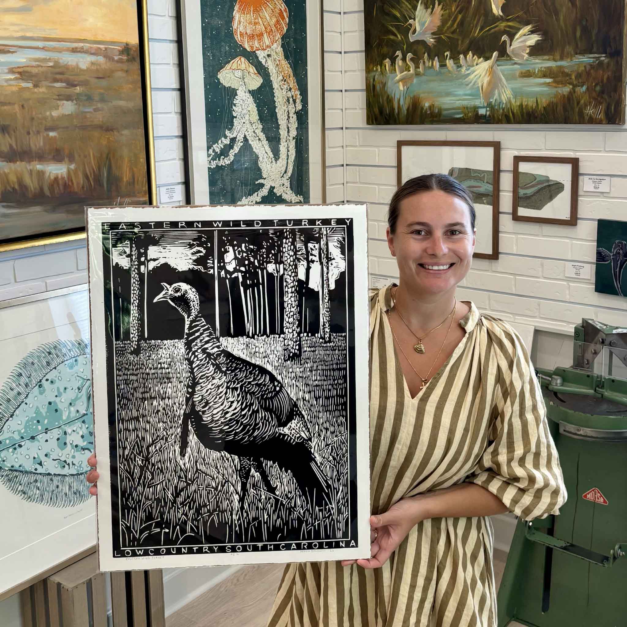 Kyra holding Eastern Wild Turkey by South Carolina artist Aaron Baldwin, a large original hand-pulled black linocut depicting a wild turkey among tall grasses and pine trees with “Lowcountry South Carolina” hand-carved along the bottom, photographed inside Gray Man Gallery in Pawleys Island.