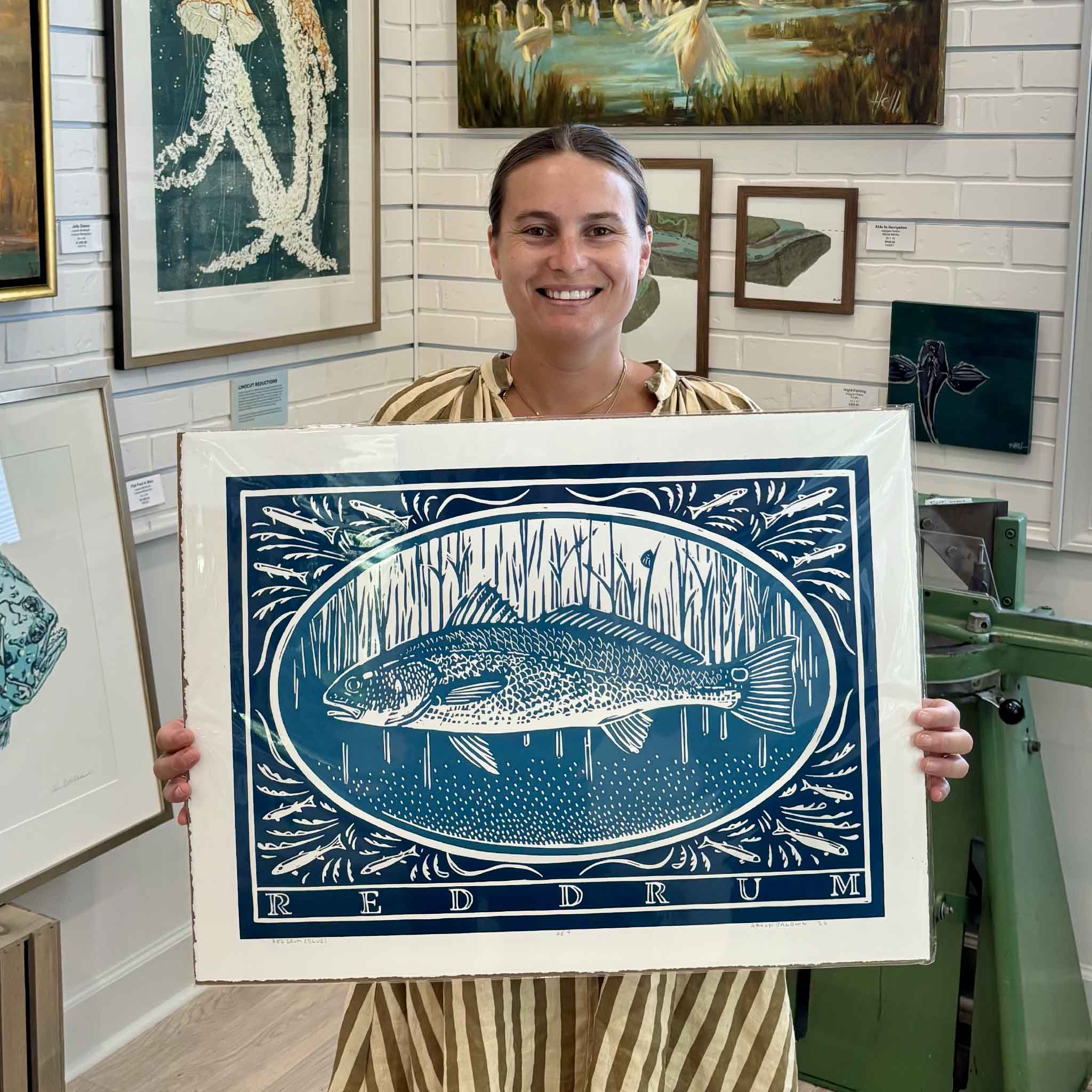 Kyra holding Red Drum by South Carolina artist Aaron Baldwin, a large original hand-pulled linocut featuring a red drum surrounded by small baitfish in a bold blue hand-carved composition, photographed inside Gray Man Gallery in Pawleys Island, South Carolina.