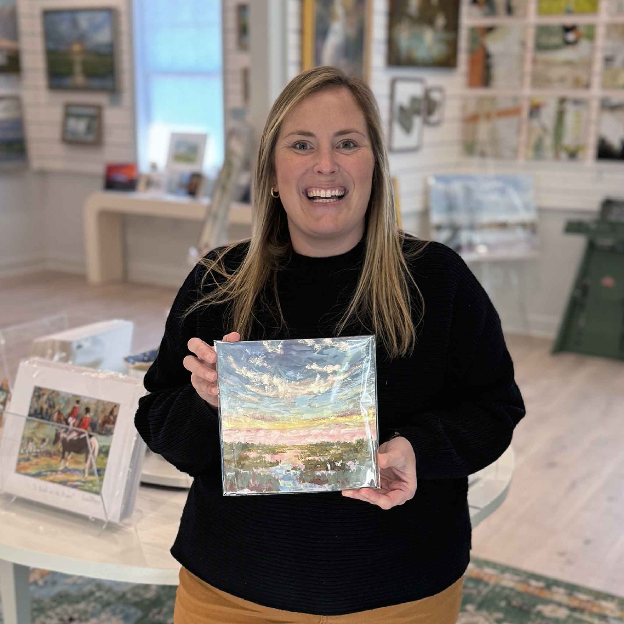 Gray Man Gallery team member holding “Afterglow Marsh,” a hand-embellished 8x8 fine art giclée with gouache by Jeanne O’Neal, showcasing original Lowcountry coastal artwork in the gallery